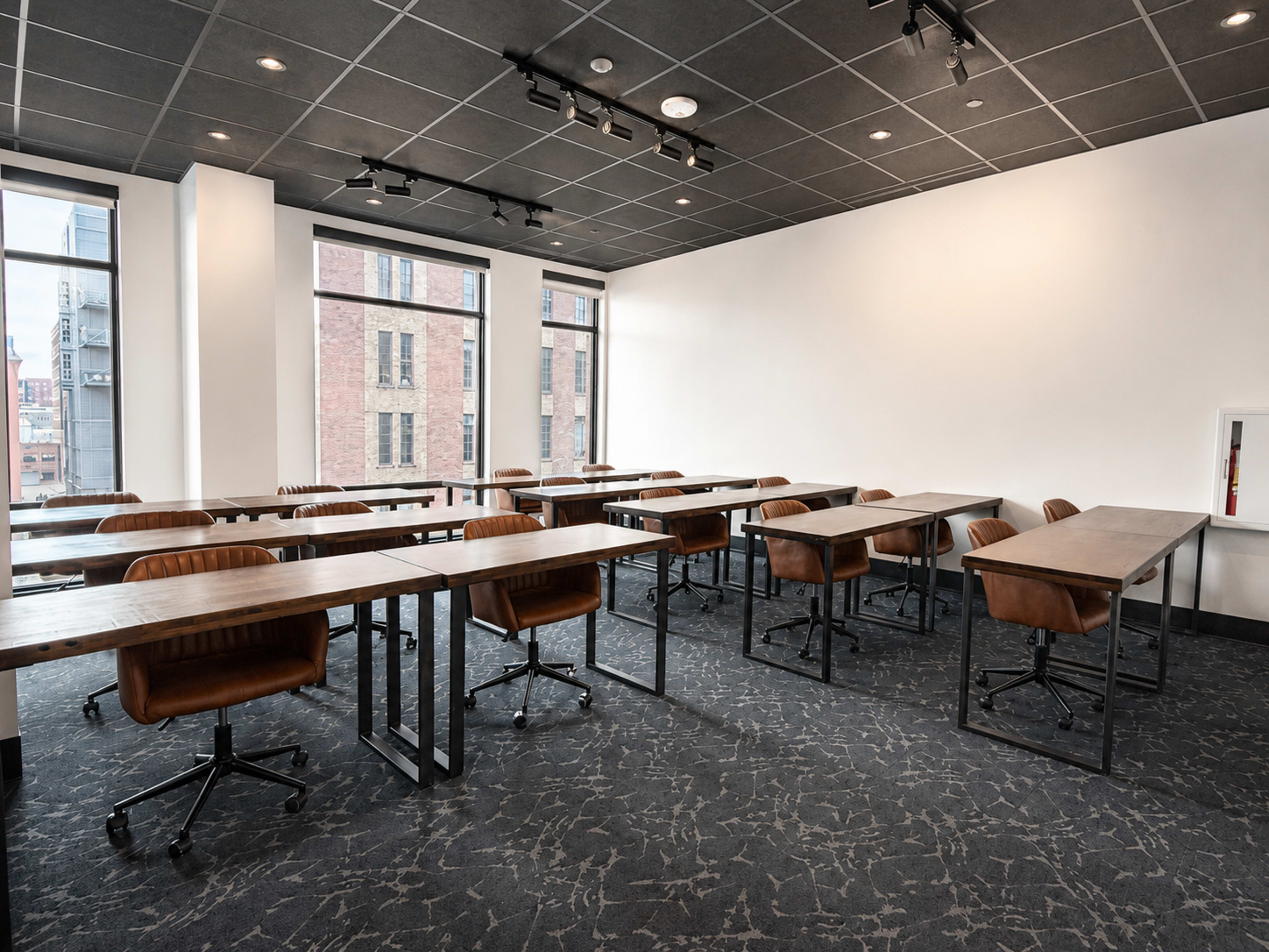 The image shows a room with several rows of tables and chairs arranged in a classroom or meeting setup, with large windows letting in natural light.