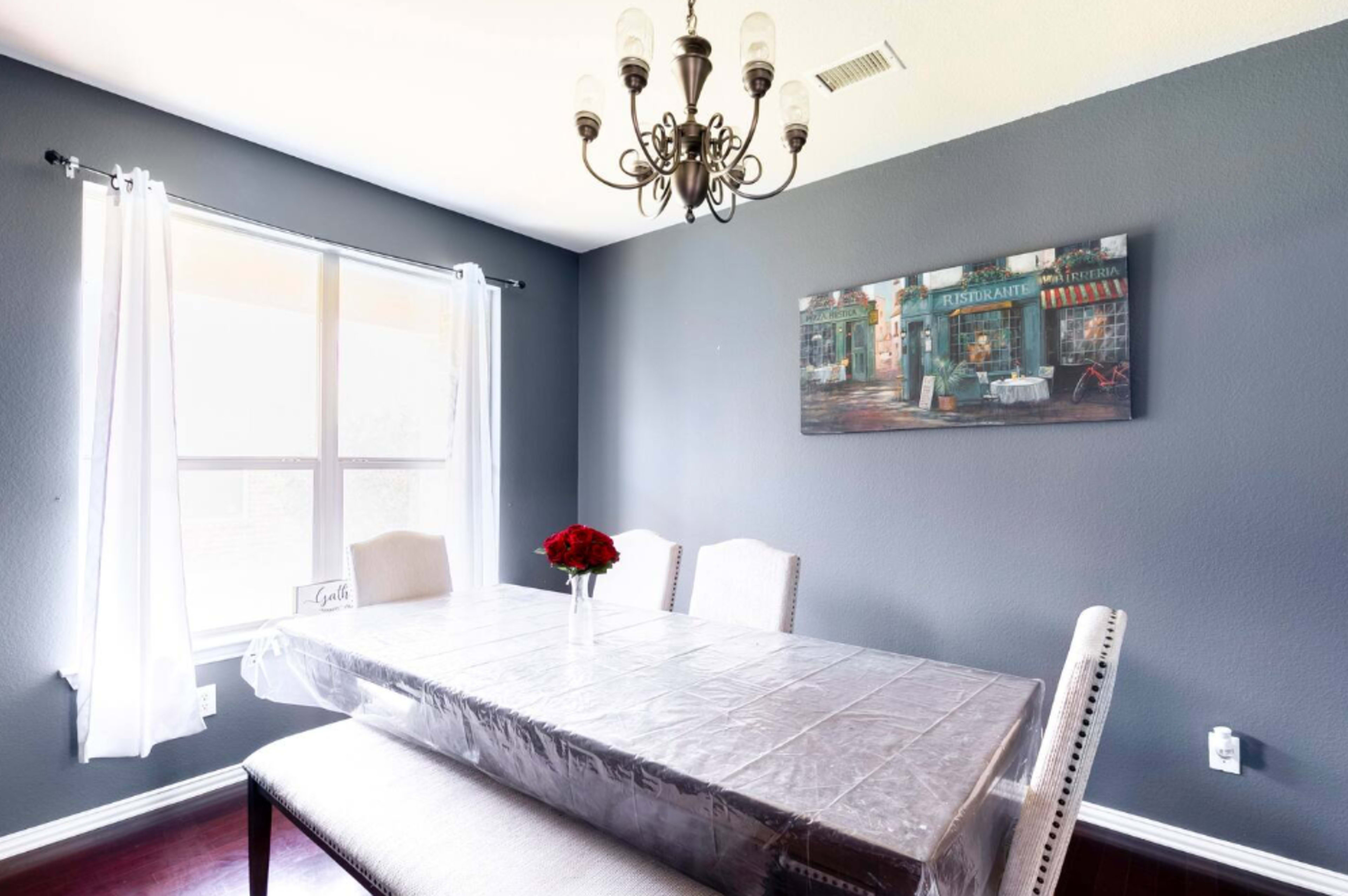 A dining room features a large table covered with a plastic cloth, surrounded by upholstered chairs, with a chandelier overhead and a painting on the wall.