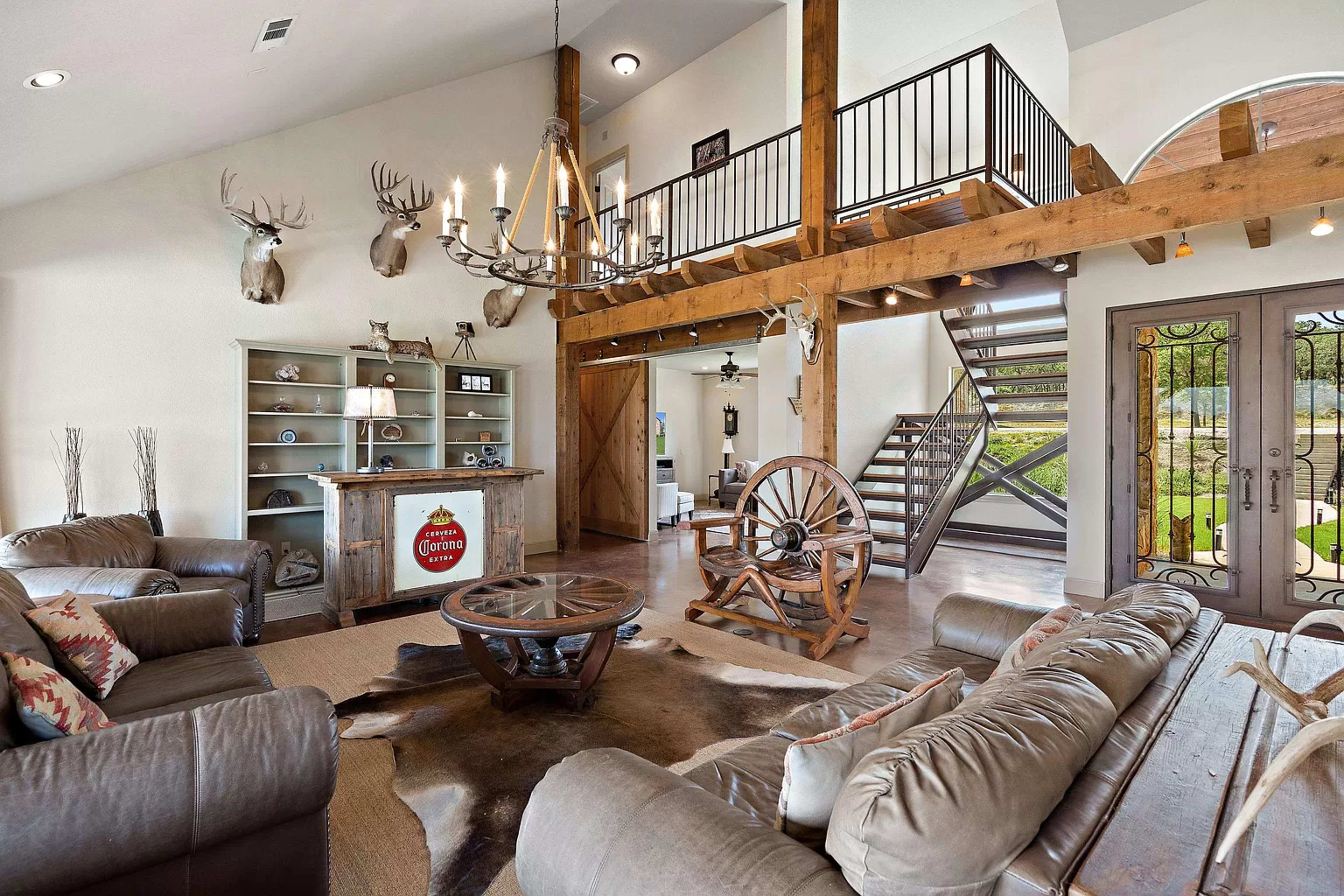 The living area features a spacious layout with a high ceiling, wooden beams, a staircase, and rustic decor, including mounted deer heads and a cowboy-themed bar.