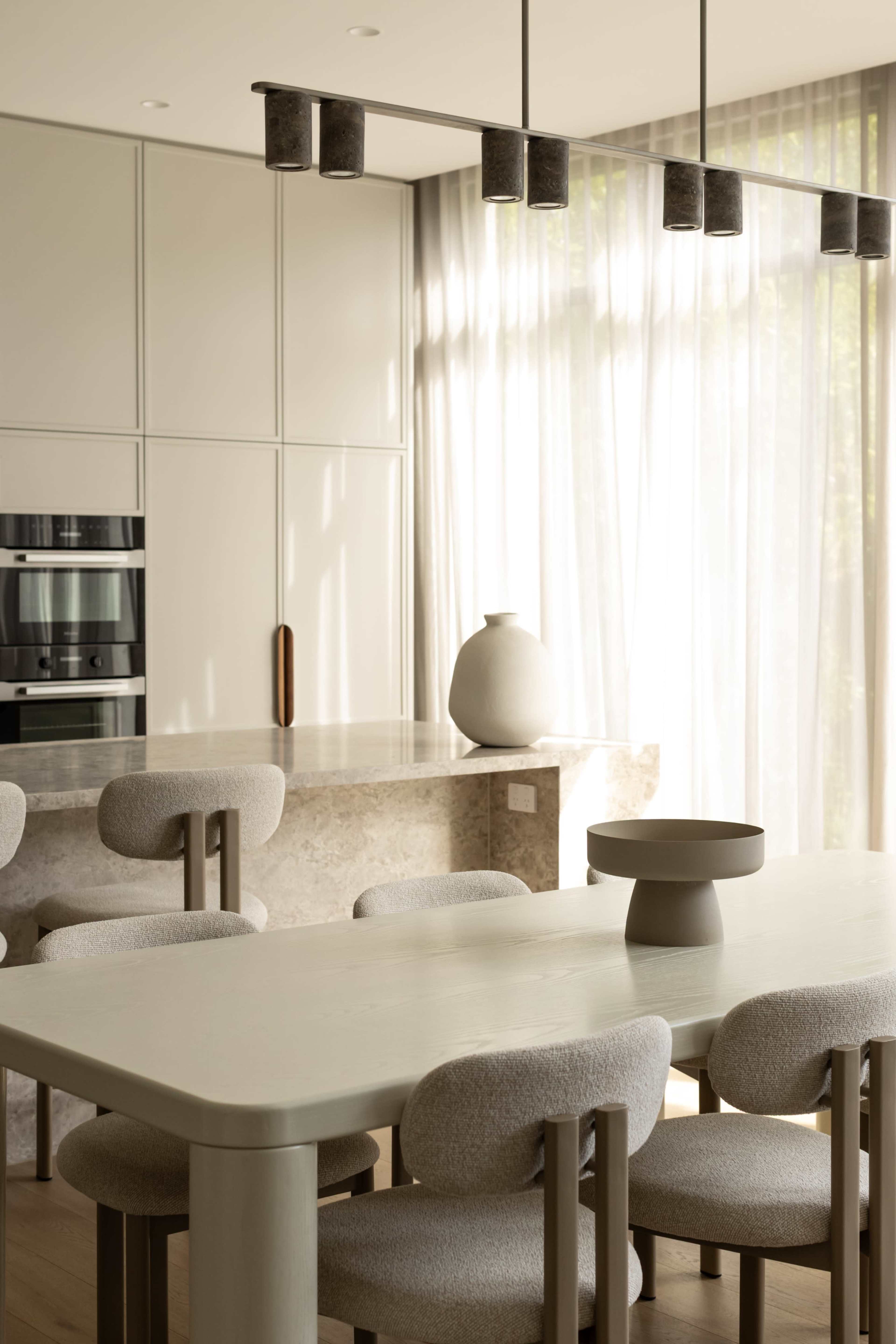 A modern kitchen features a light-colored table surrounded by rounded chairs, with a countertop, stylish pendant lights, and large windows allowing natural light to enter.