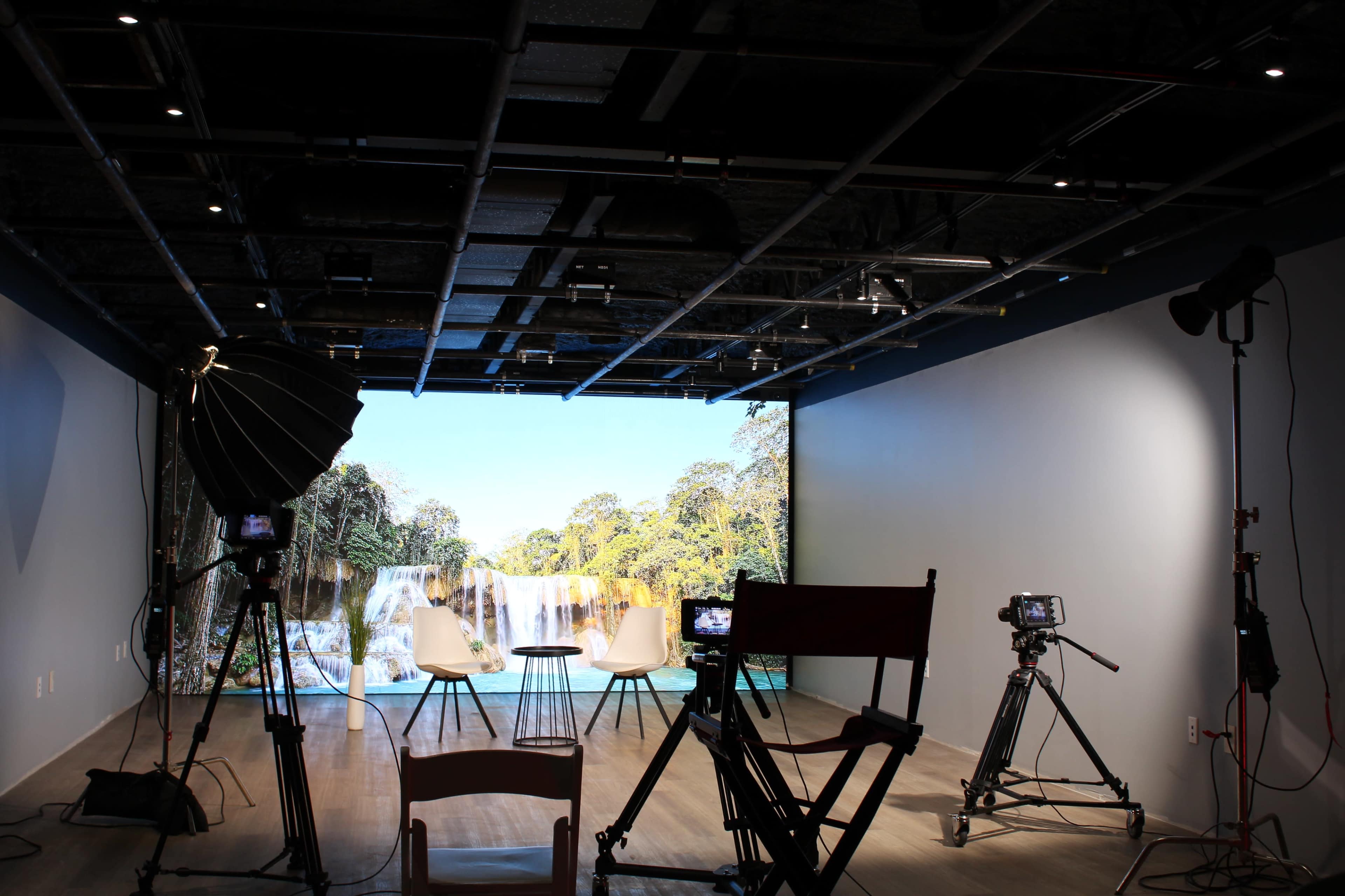The image shows a studio setup with two white chairs facing a large backdrop featuring a waterfall, surrounded by various filming equipment and lighting.