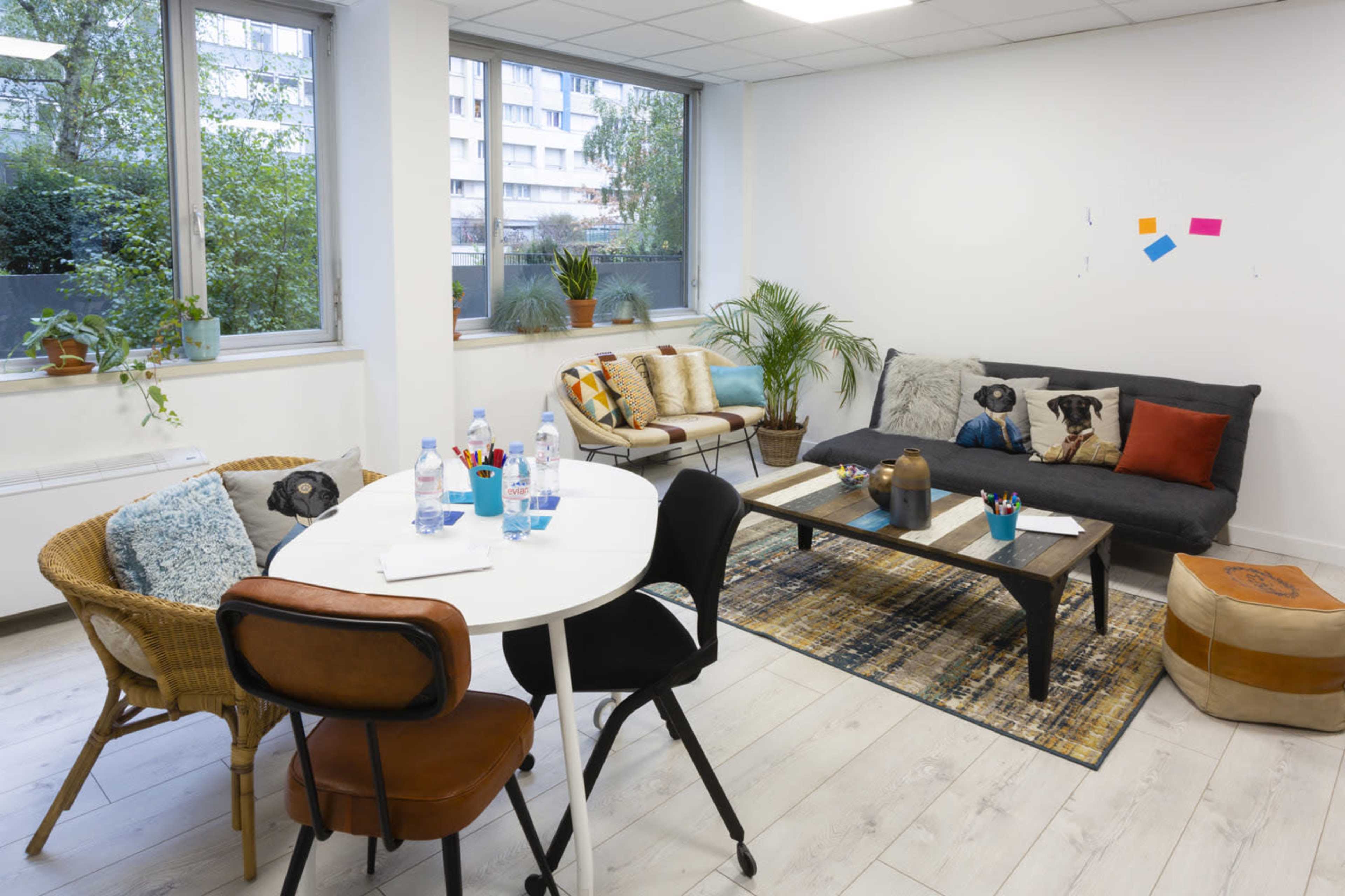 The image shows a bright office lounge area with a black sofa, a round white dining table with chairs, and several potted plants.