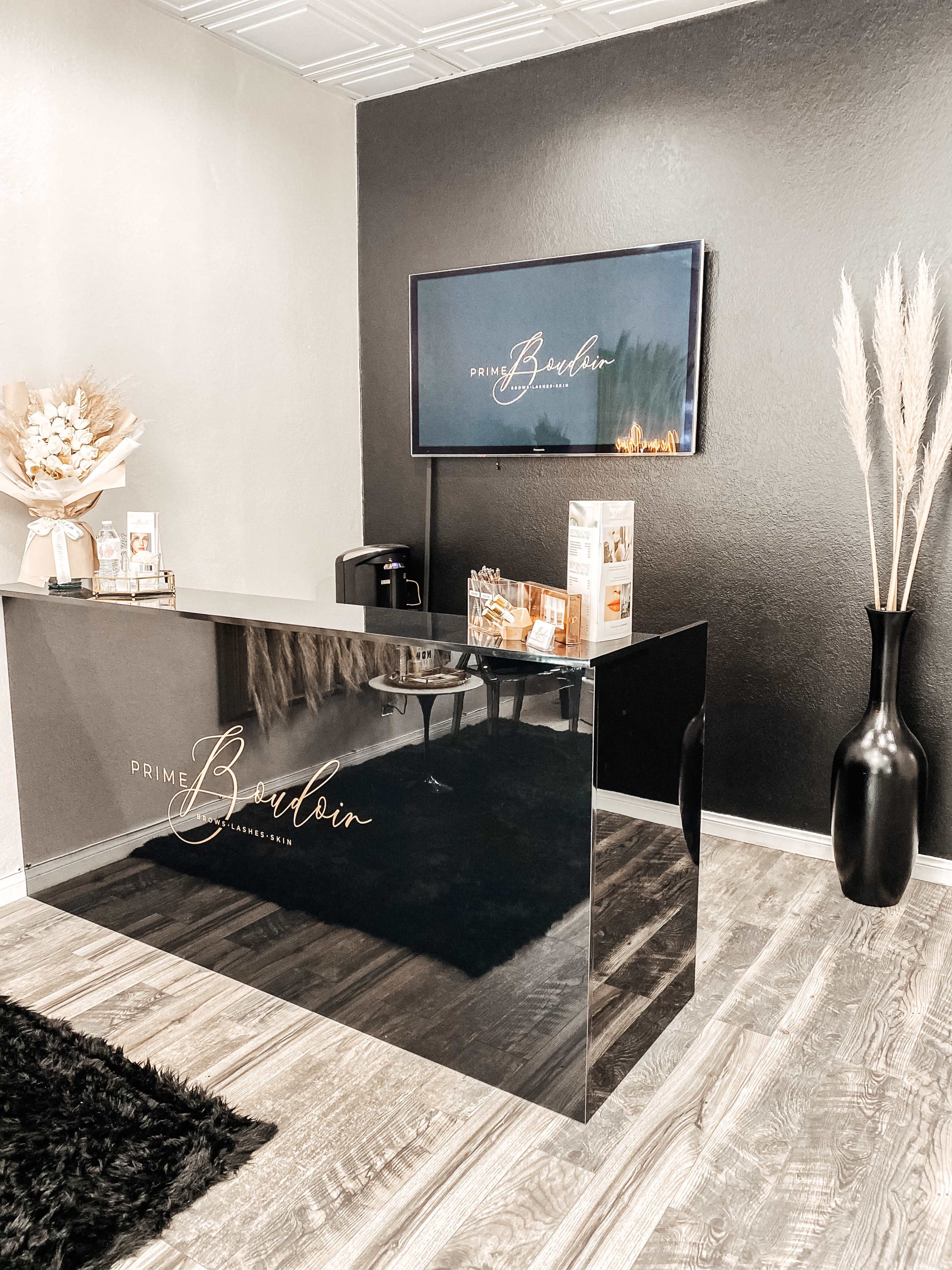 A modern reception area with a sleek, dark-colored front desk, a wall-mounted television displaying a logo, and decorative elements such as a large vase and pampas grass.