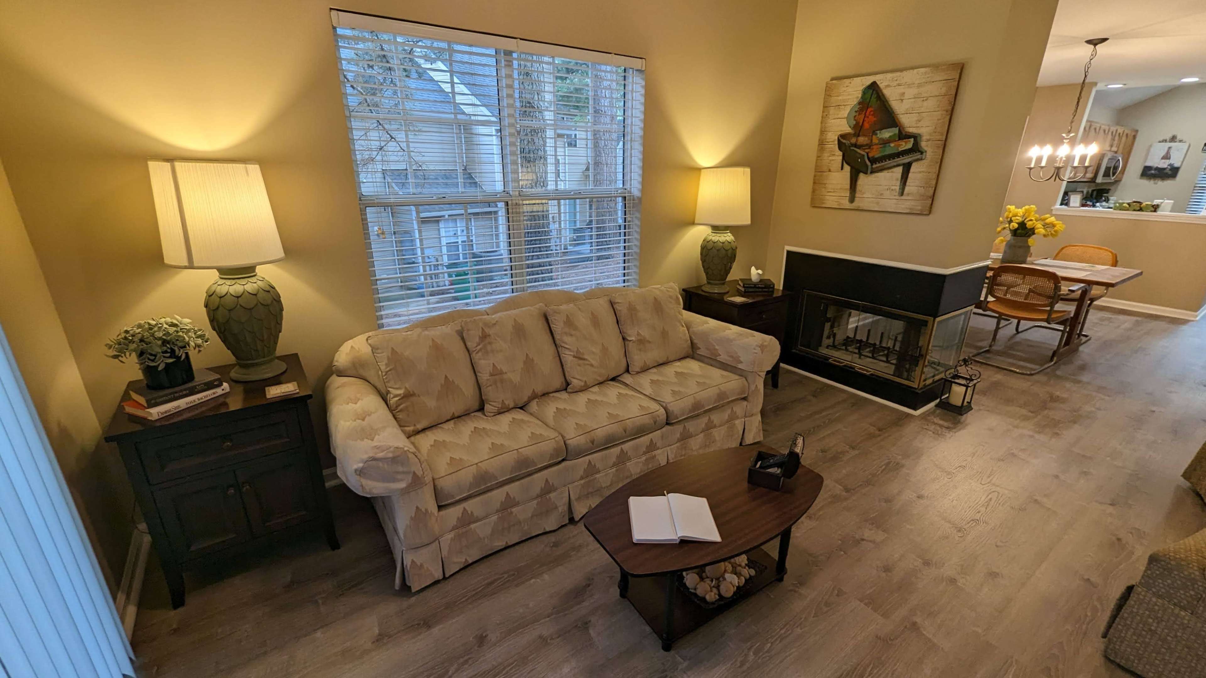 The image shows a living room with a beige couch, two lamps, a coffee table with a notebook, and a fireplace, alongside a dining area visible in the background.