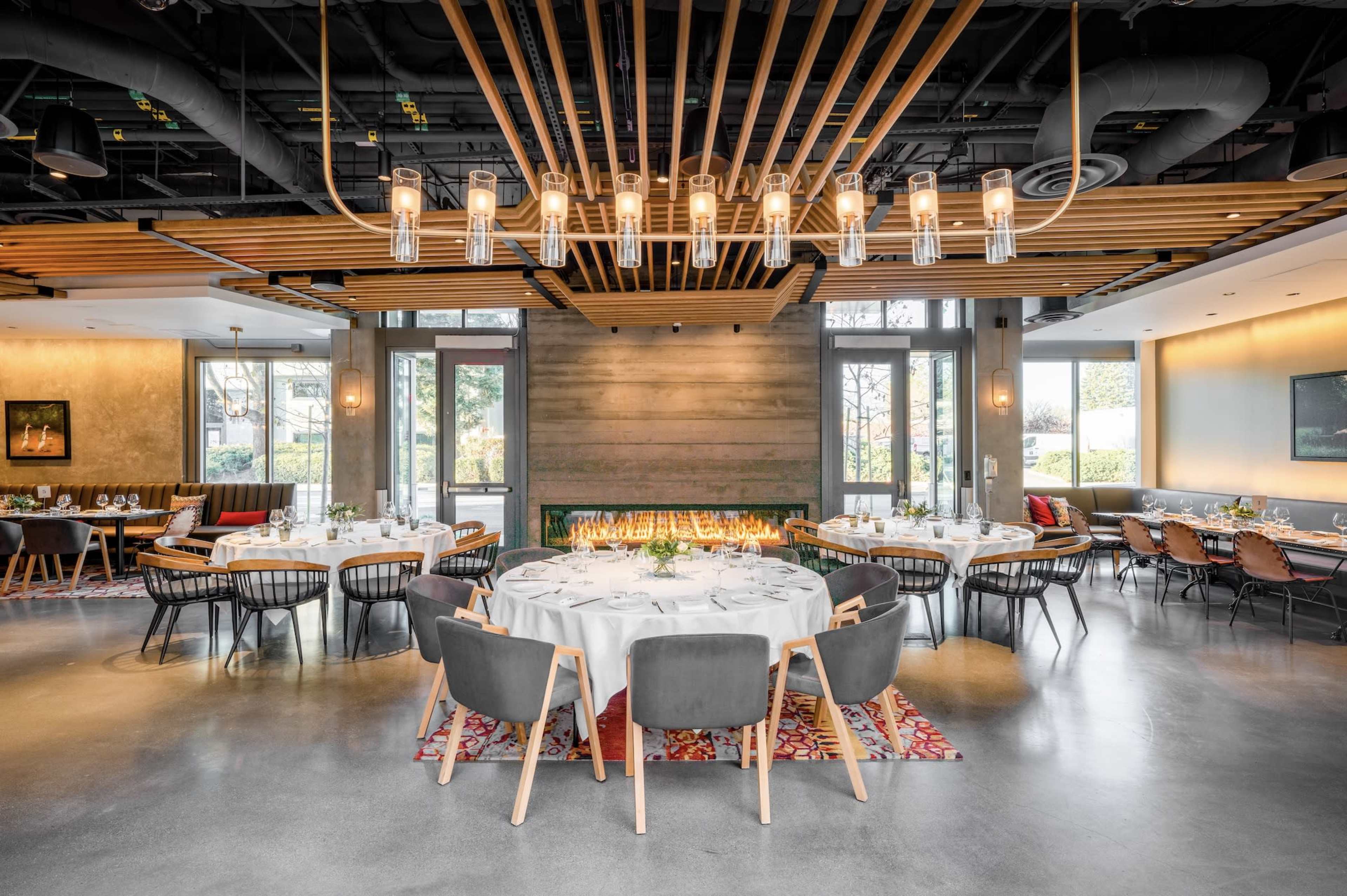 A modern restaurant interior features round tables set with white tablecloths and stylish chairs, illuminated by pendant lights and a linear wood ceiling design.