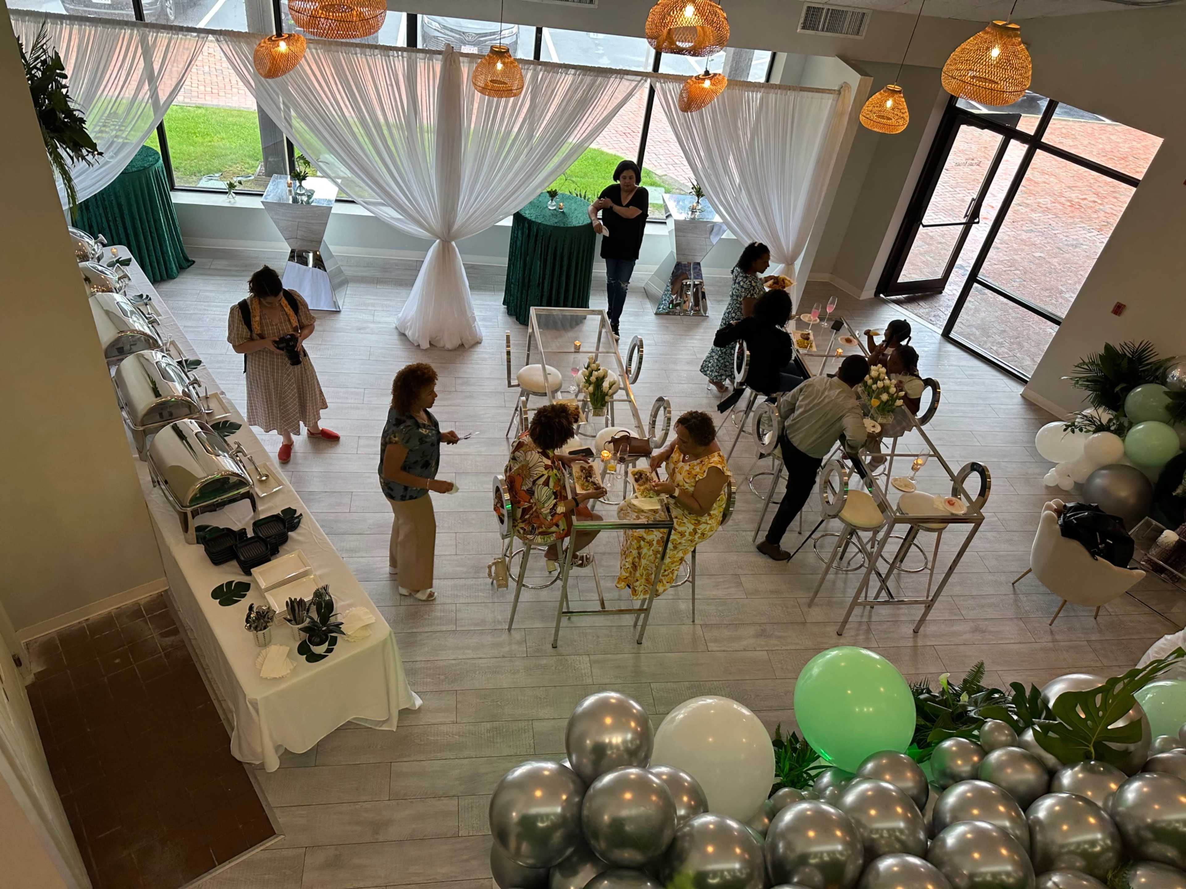 A bright event space with tables set for dining, guests gathering, and decorative elements like balloons and hanging lights.