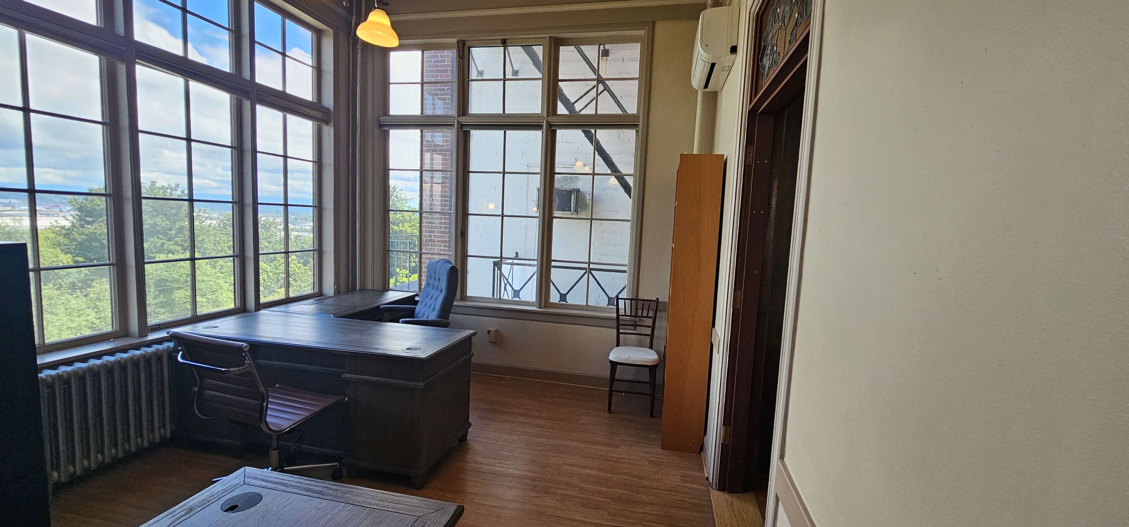 The image shows a bright office space with large windows, a wooden desk and chair, and a white chair positioned against the wall.