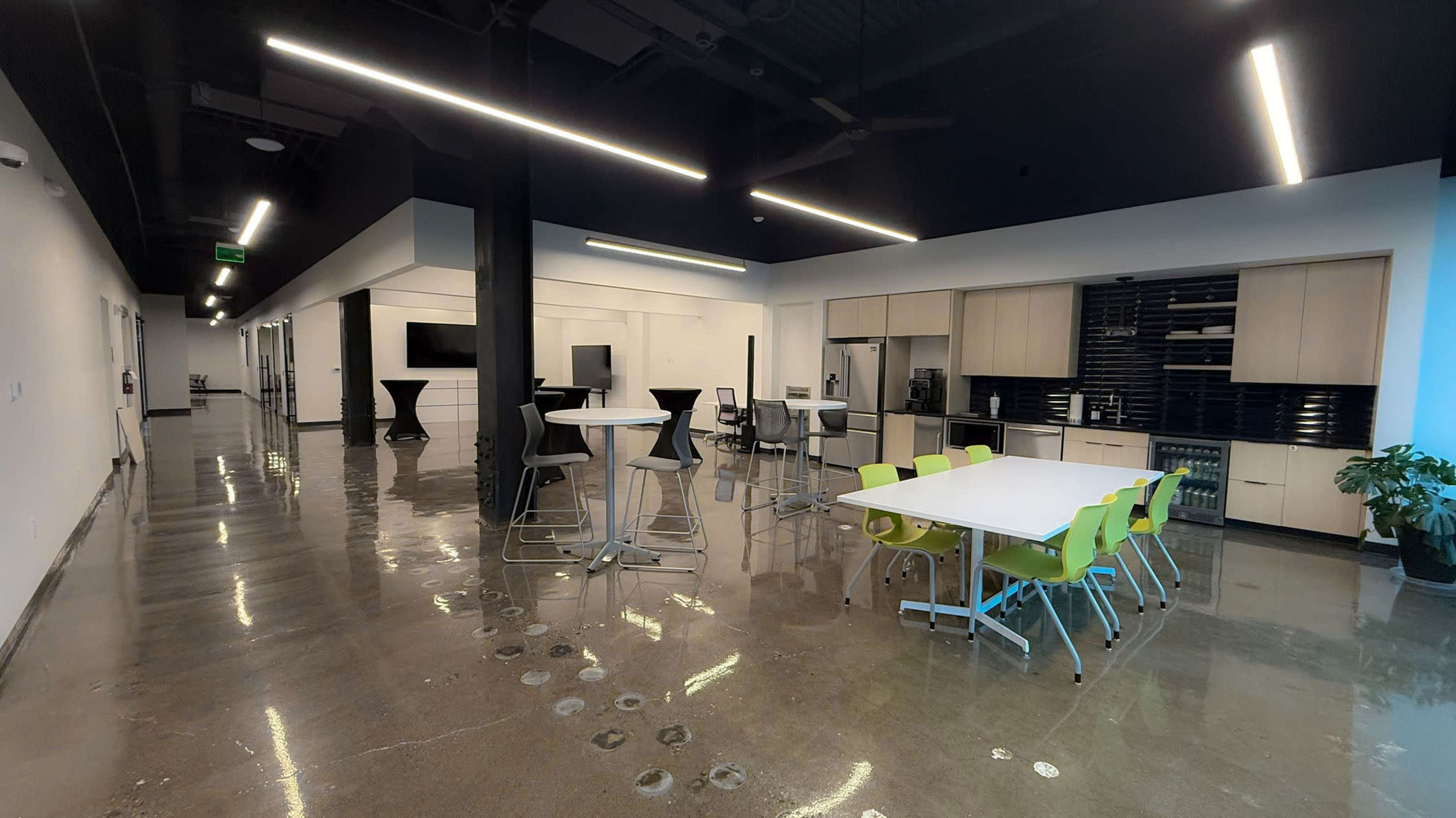 The image shows a modern office break room with a polished concrete floor, a kitchen area, various seating options, and bright fluorescent lighting.