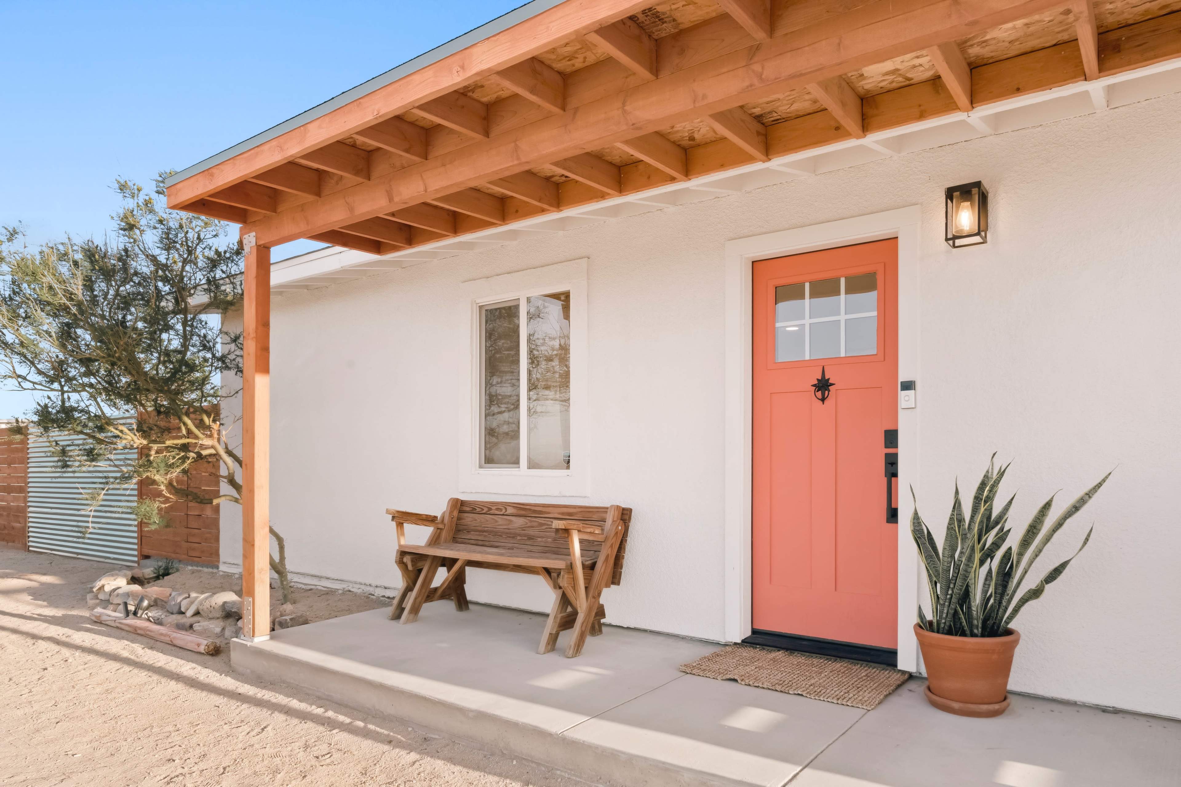 A house exterior featuring a coral-colored front door, a wooden bench, and a potted plant on a concrete patio.