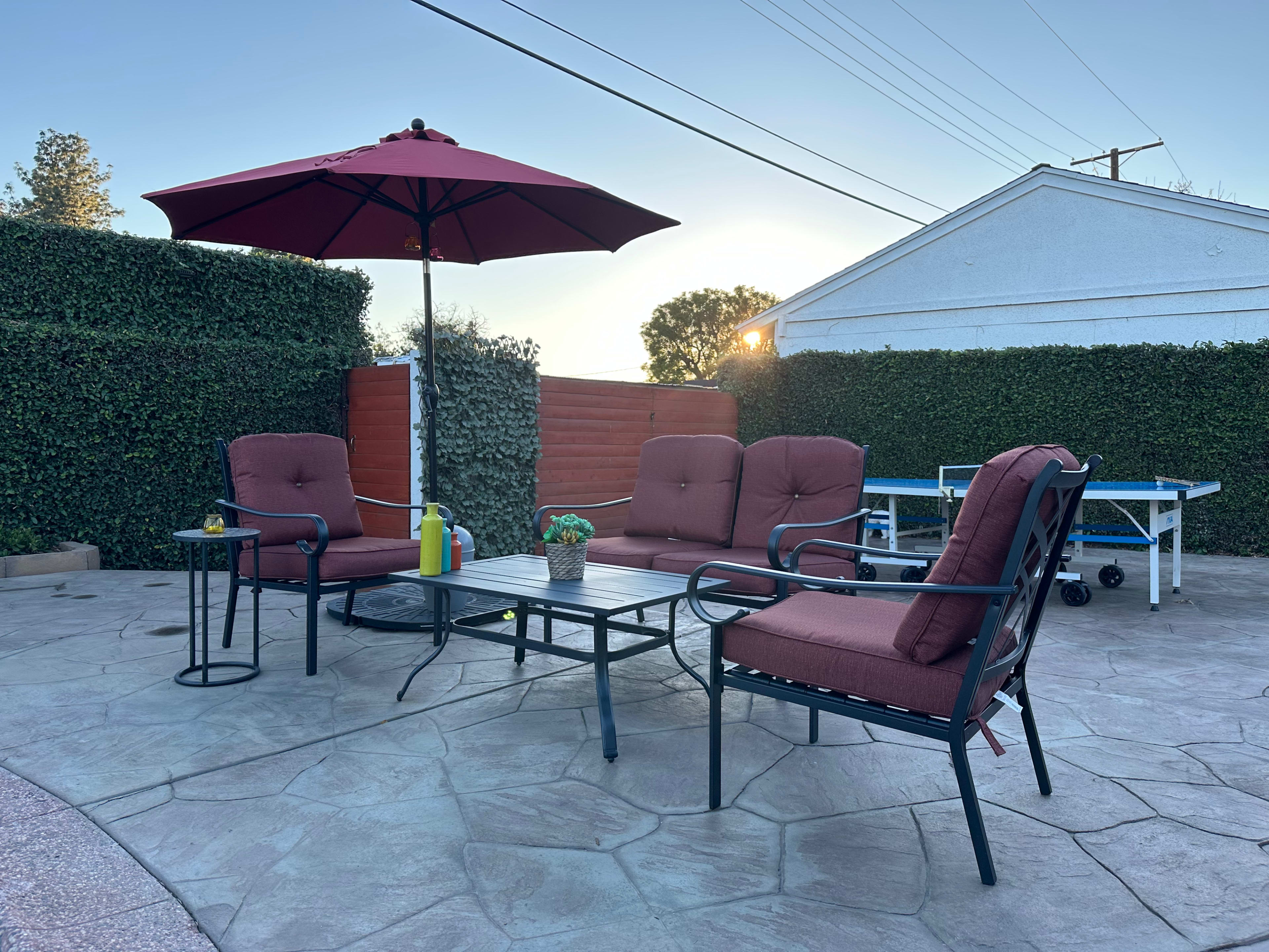 A patio area features four cushioned chairs, a table, and a large umbrella surrounded by green hedges.