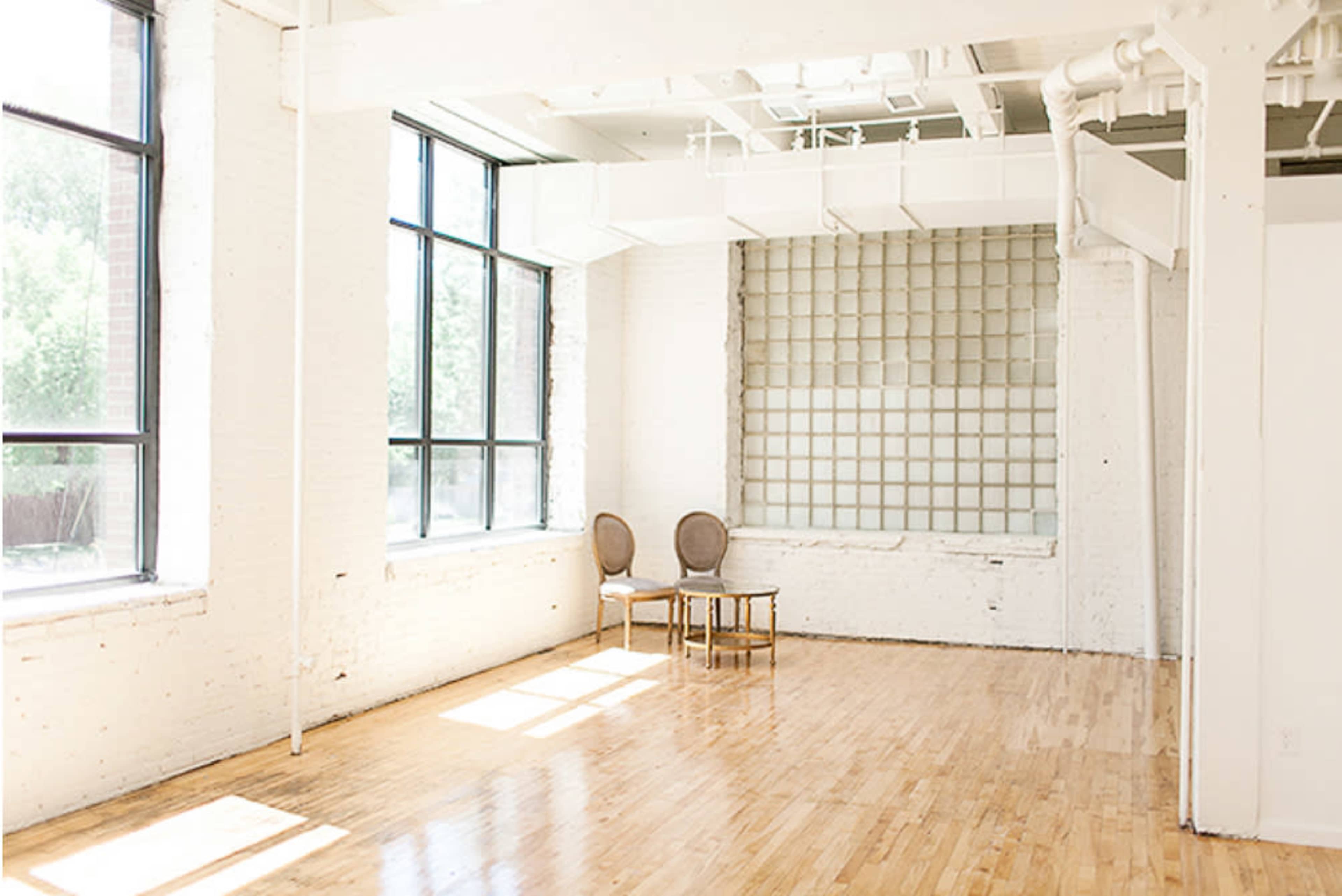 The image shows a bright, empty room with hardwood floors, large windows, and a small table with two chairs against a wall featuring a grid pattern.