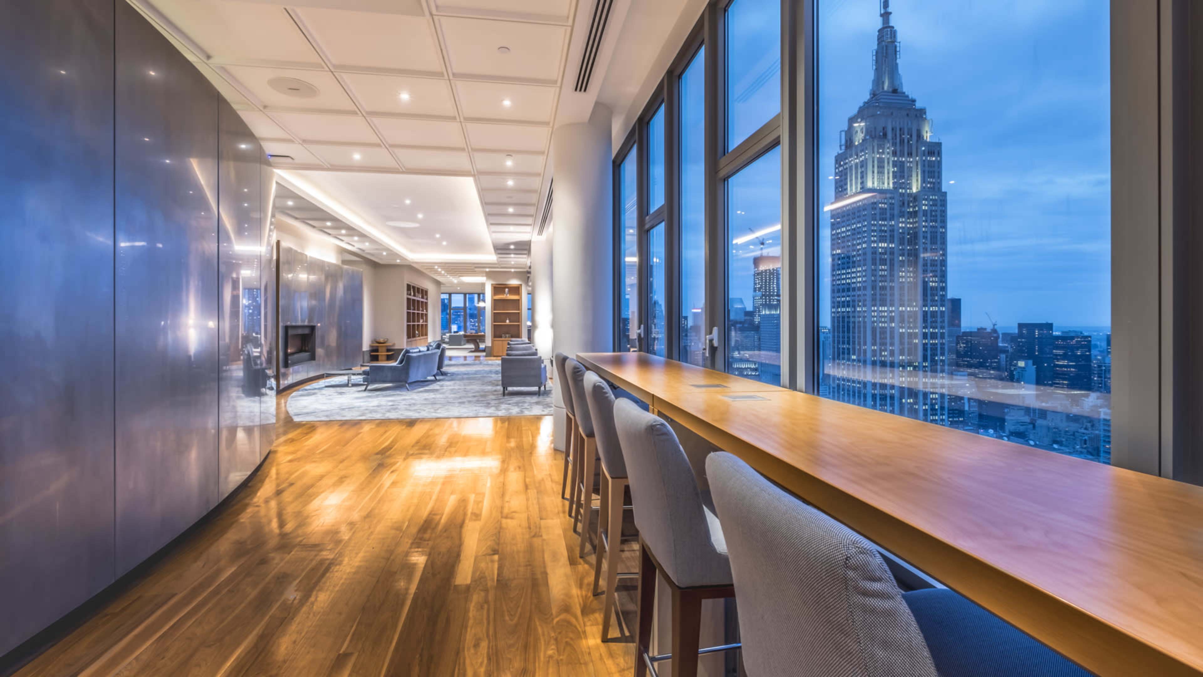 A spacious, modern interior features a long wooden bar overlooking the Empire State Building through large windows.