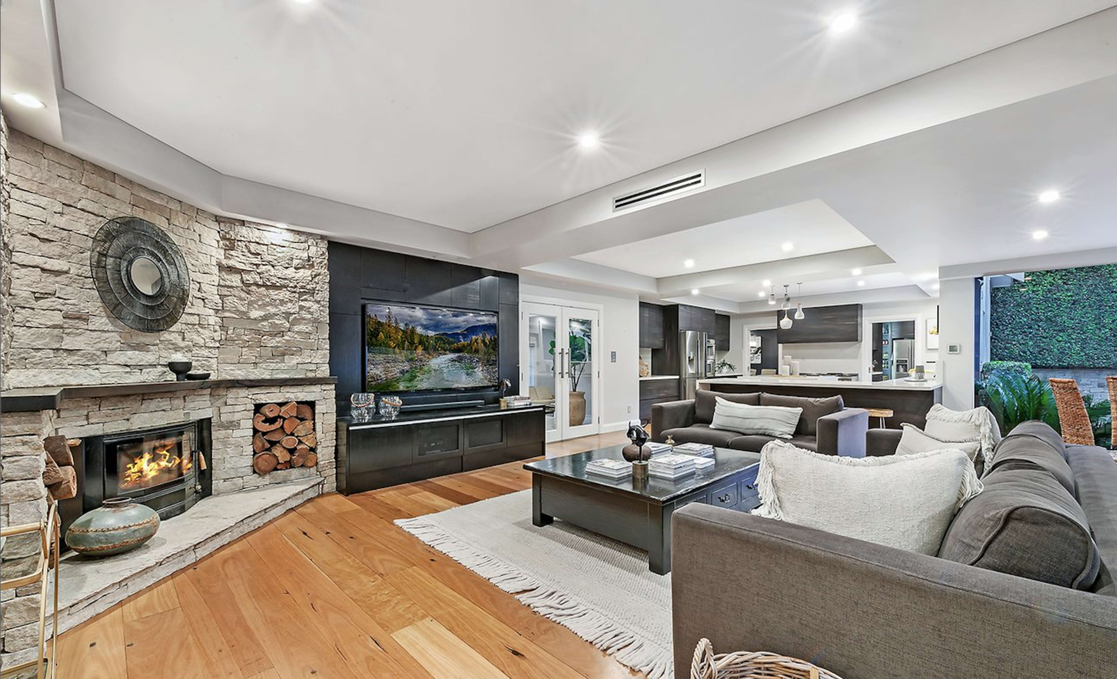 The image shows a modern living room featuring a stone fireplace, a large television on a dark cabinet, and a sectional sofa arranged around a coffee table.