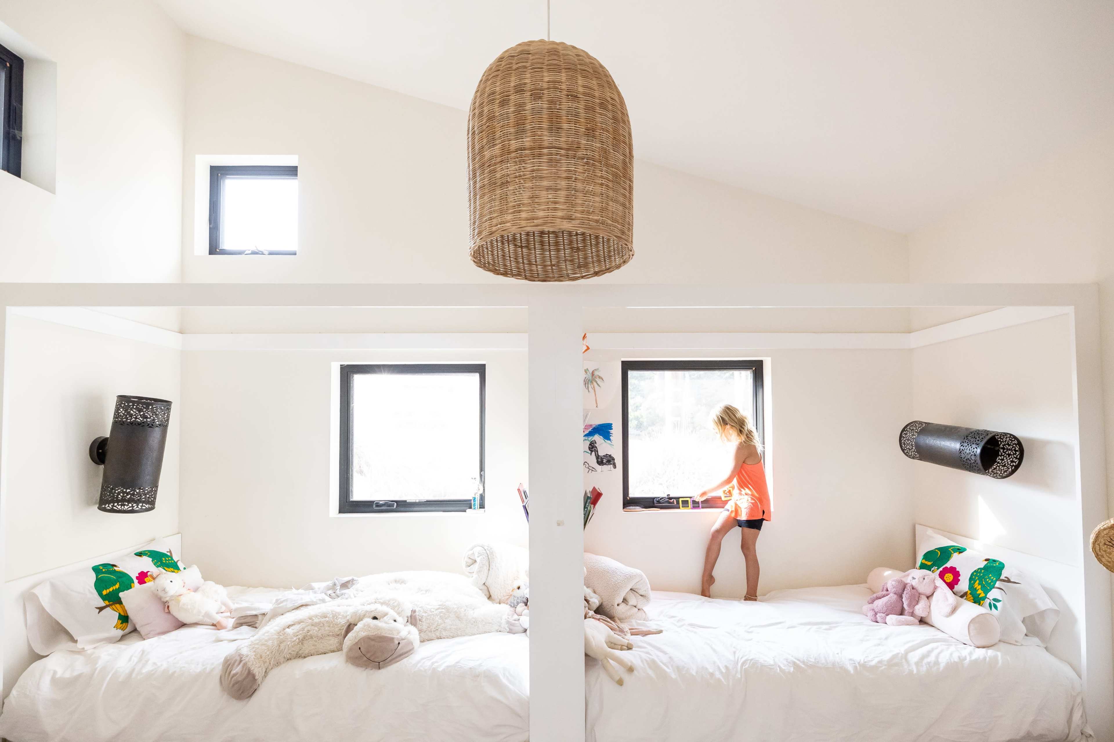 A young child stands on the edge of a bed in a brightly lit room with two separate sleeping areas adorned with stuffed animals.