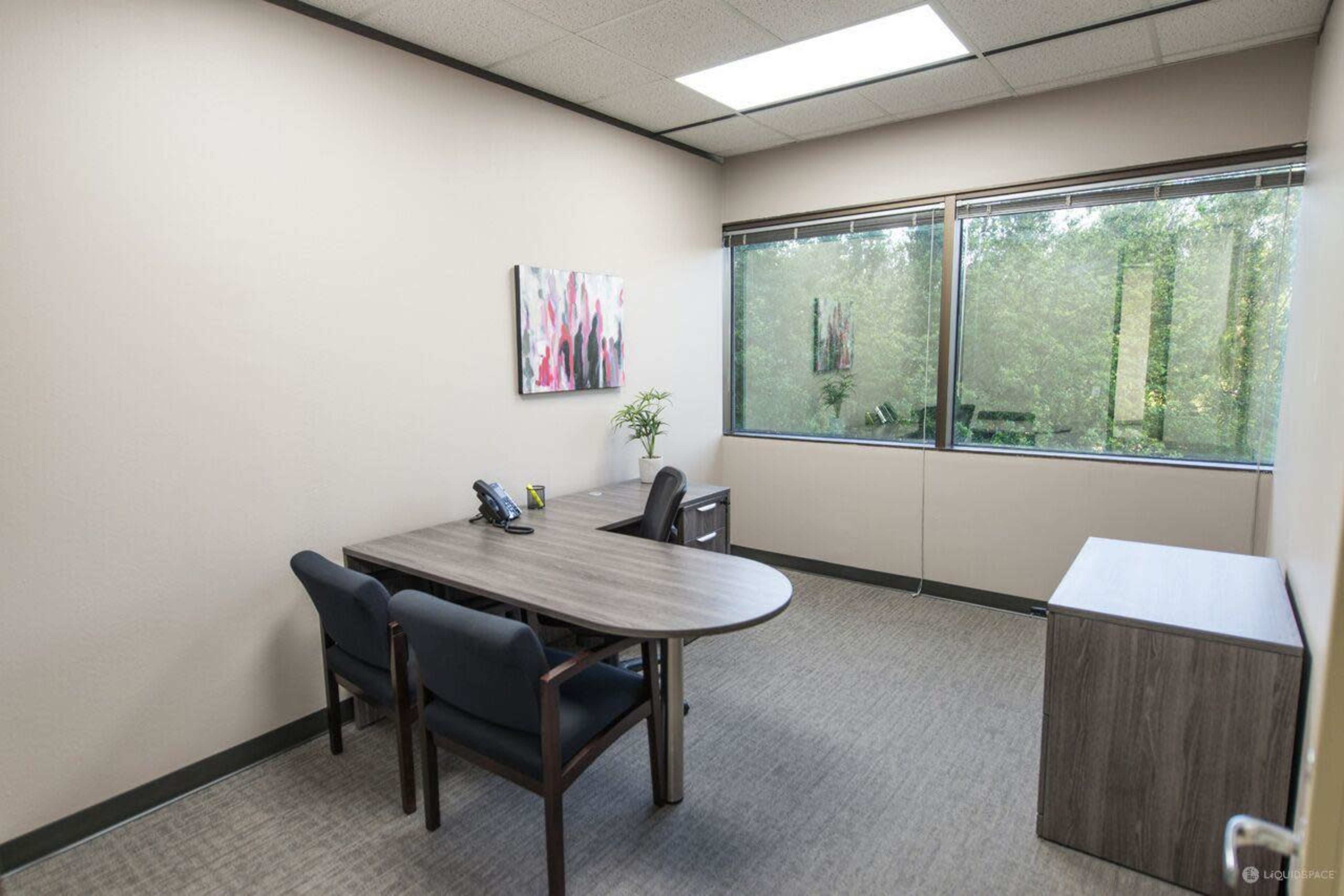 The image shows a small office with a round table, two chairs, a phone, and a window overlooking trees.