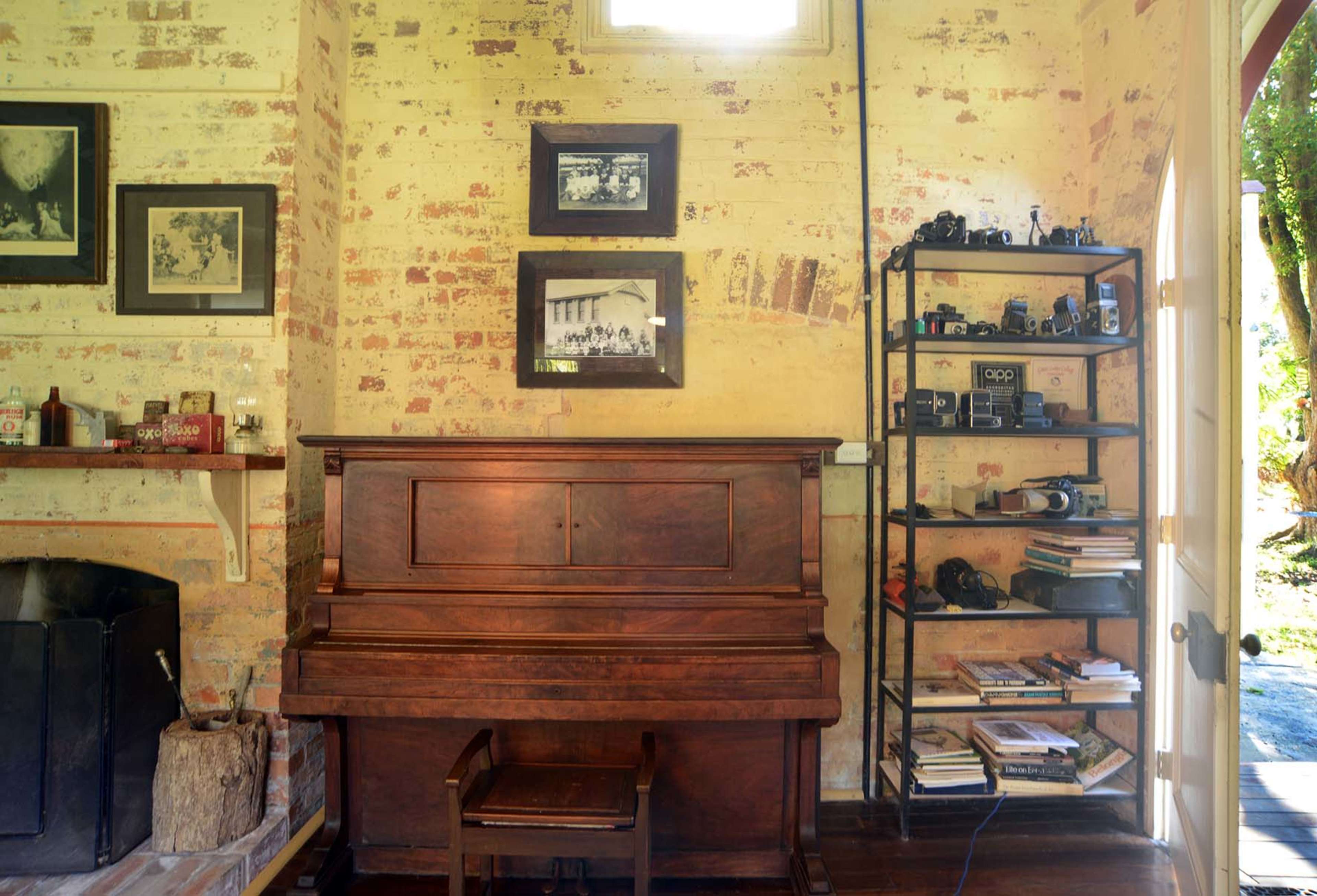 A wooden piano sits against a wall adorned with framed photographs, beside a metal shelf filled with books and cameras.