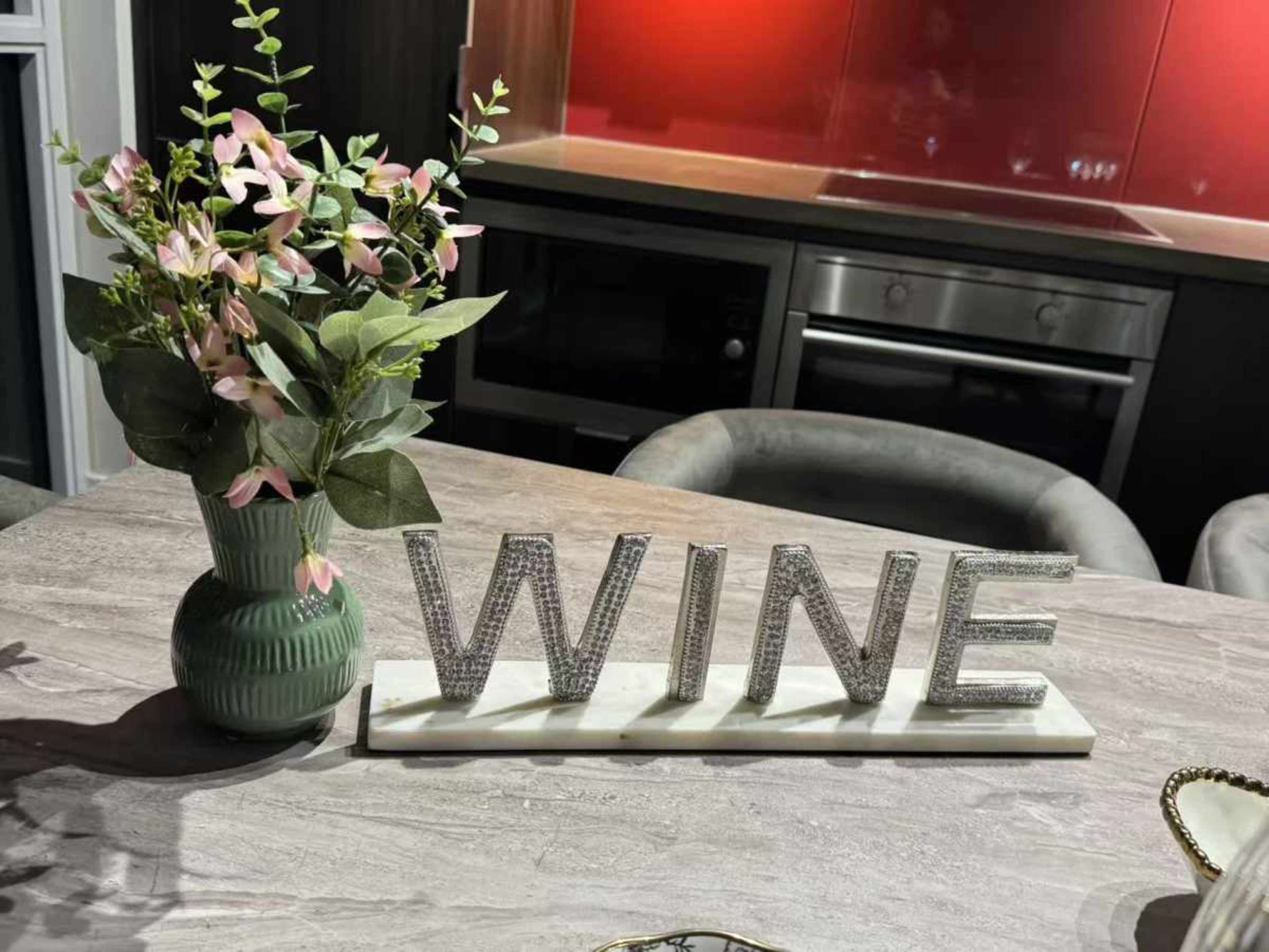 A decorative arrangement featuring a vase of flowers and a sparkling "WINE" sign sits on a marble table in a modern kitchen.