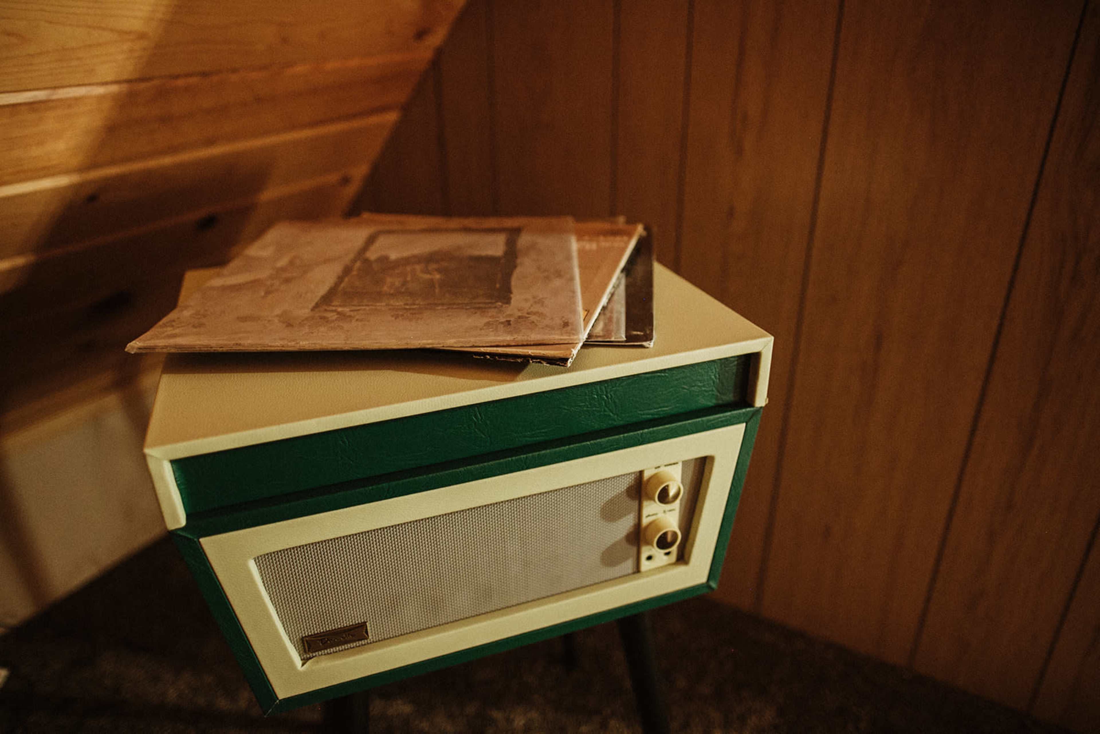 A green and cream record player sits on a small table with stacked vinyl records beside it, against a wooden wall.