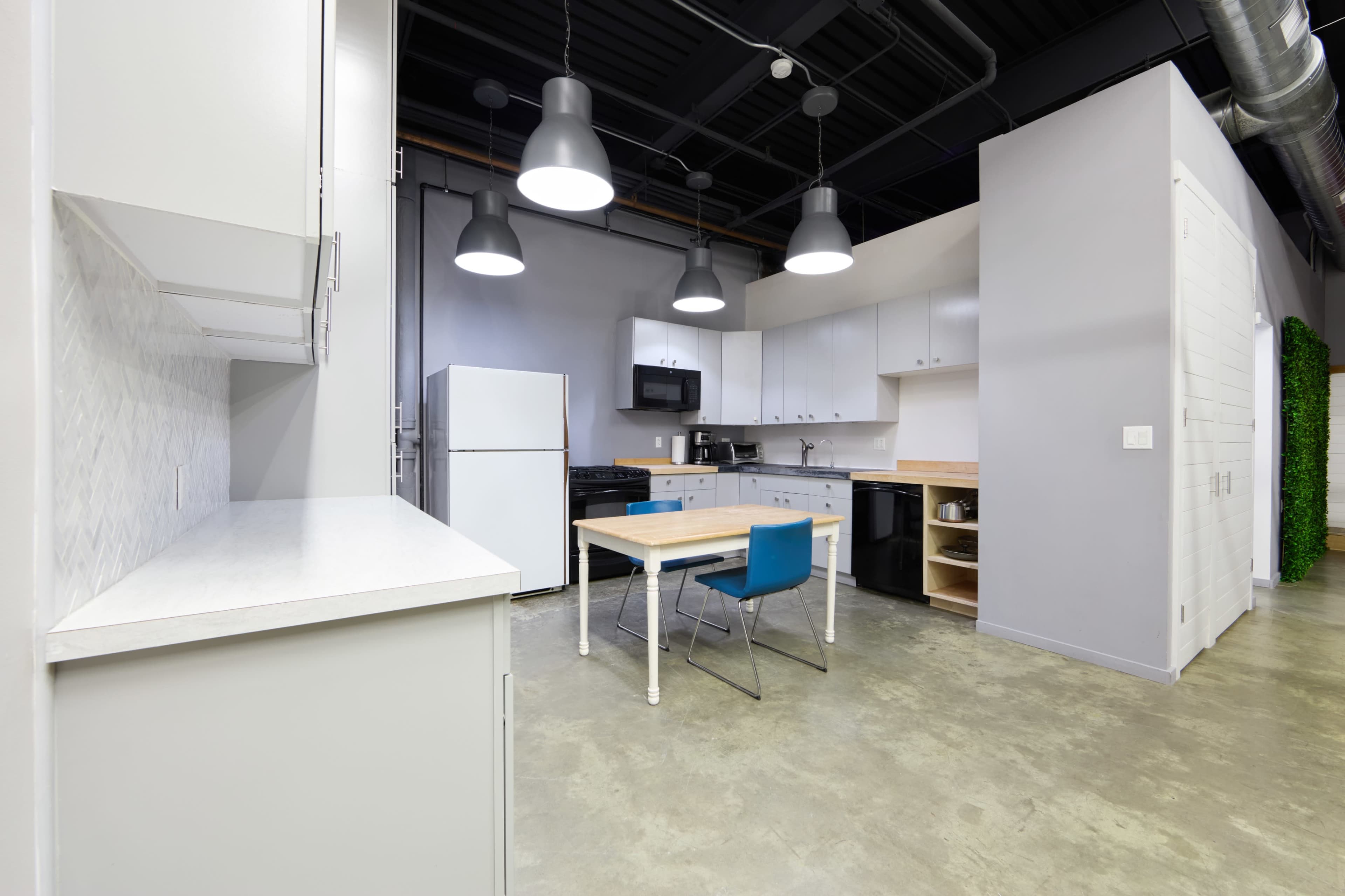 The image shows a modern kitchen with white cabinets, a dining table, blue seating, and various kitchen appliances including a refrigerator and stove.