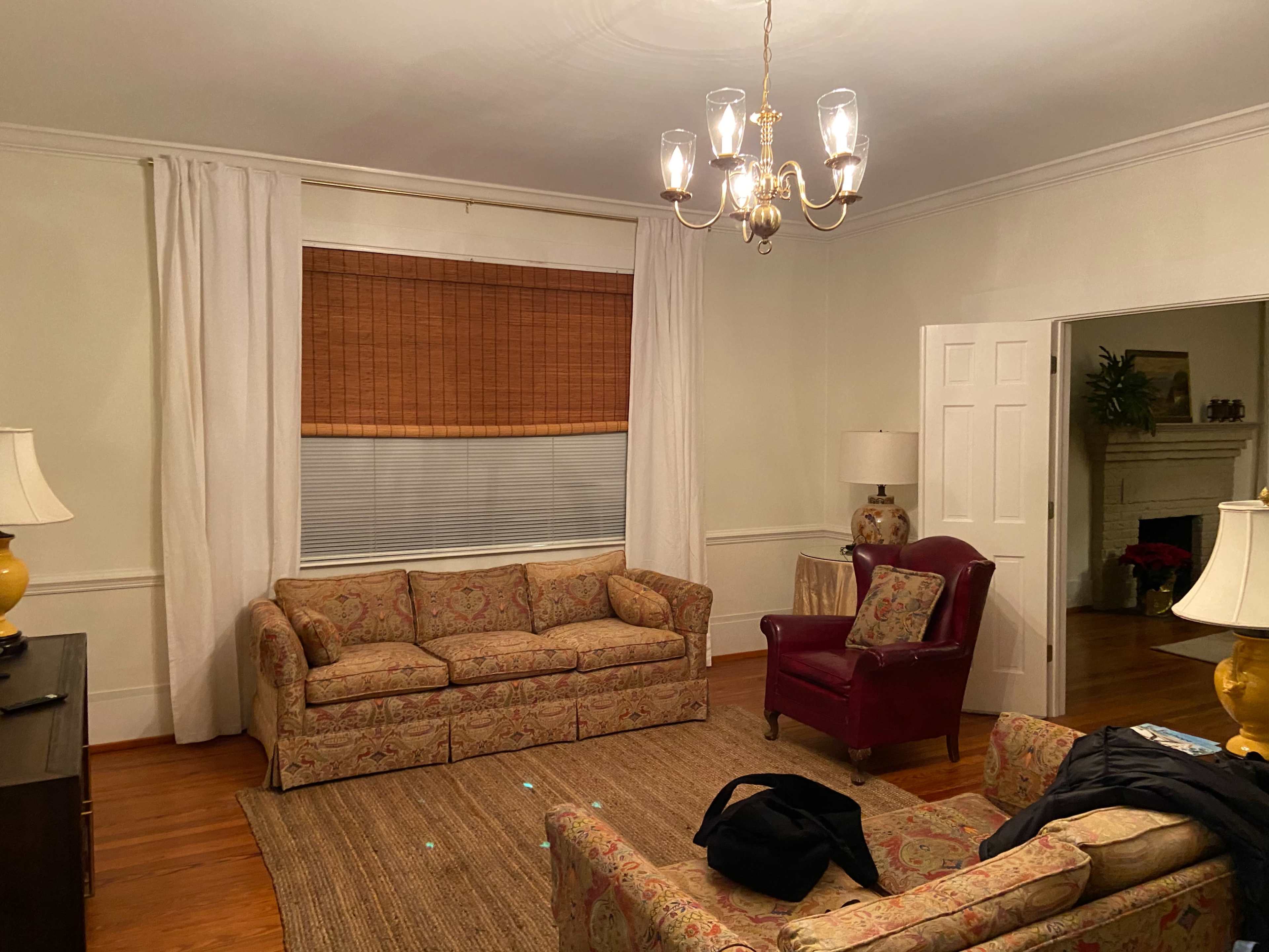 A living room with two patterned sofas, a maroon armchair, a chandelier, and a window covered with bamboo shades.