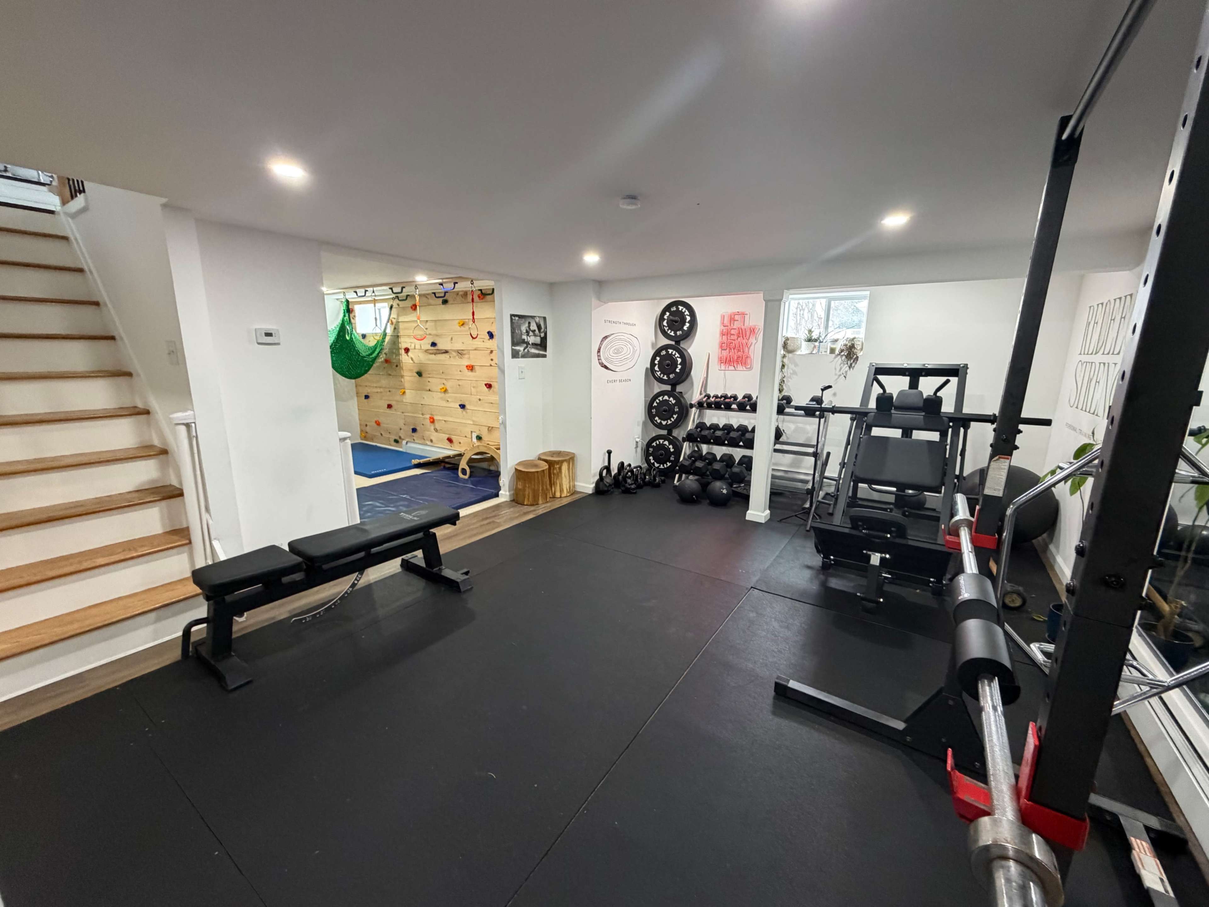 The image shows a home gym with various exercise equipment, including free weights, a bench, a squat rack, and a section for climbing.