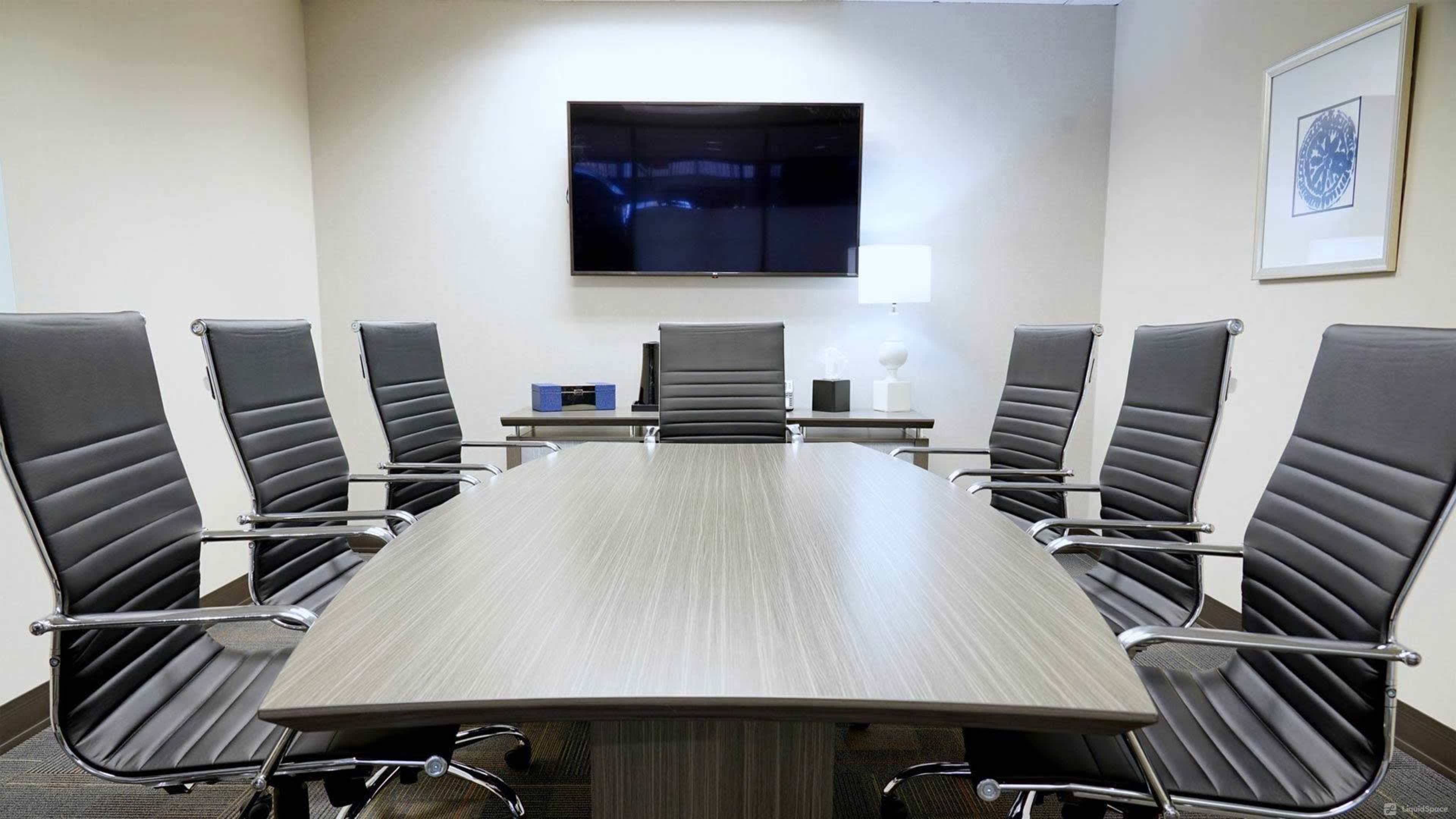 A modern conference room features a large table surrounded by six sleek chairs, with a television mounted on the wall and a lamp on the side.
