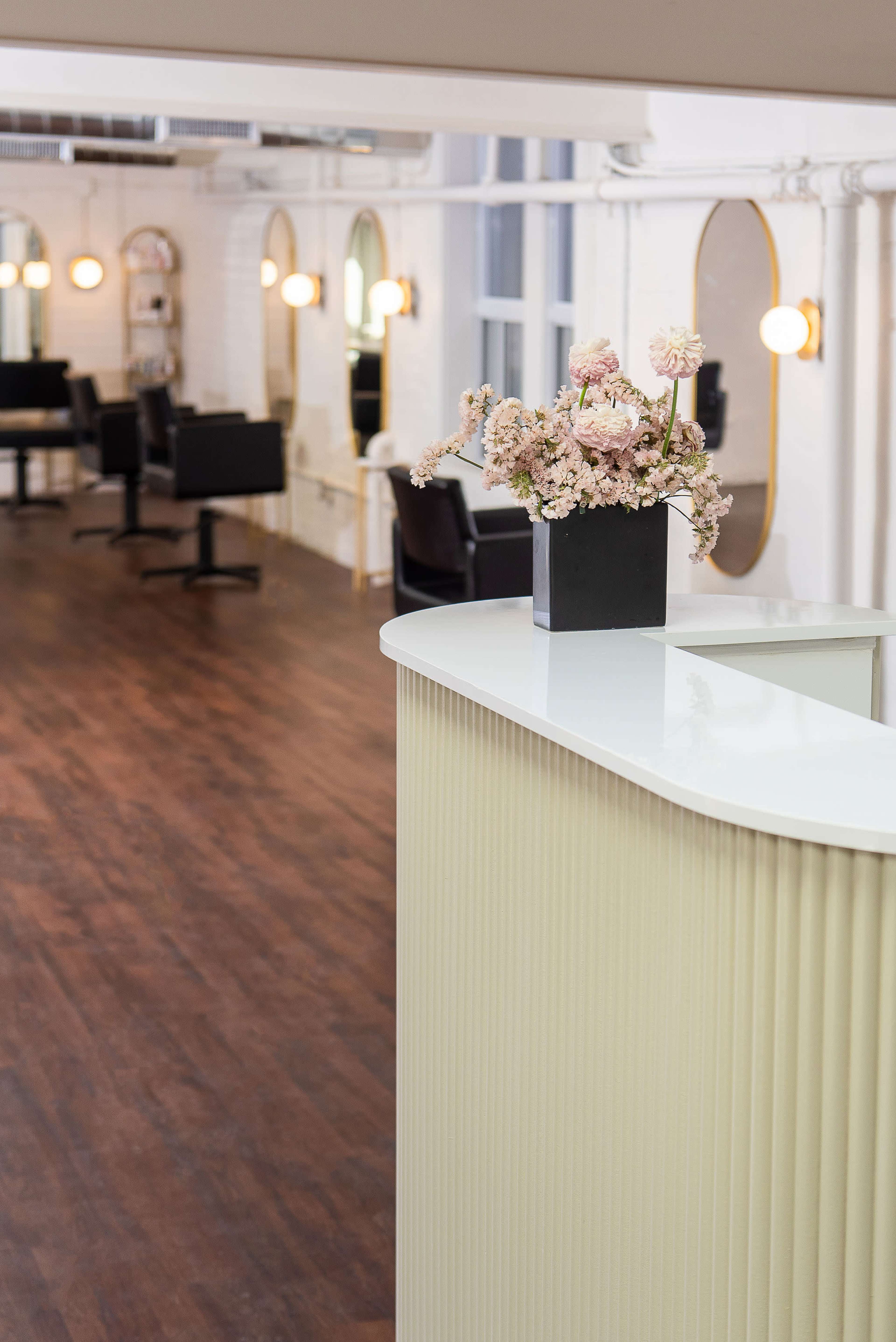 A modern interior of a salon features a reception desk with a vase of flowers, wooden flooring, and illuminated mirrors along the walls.