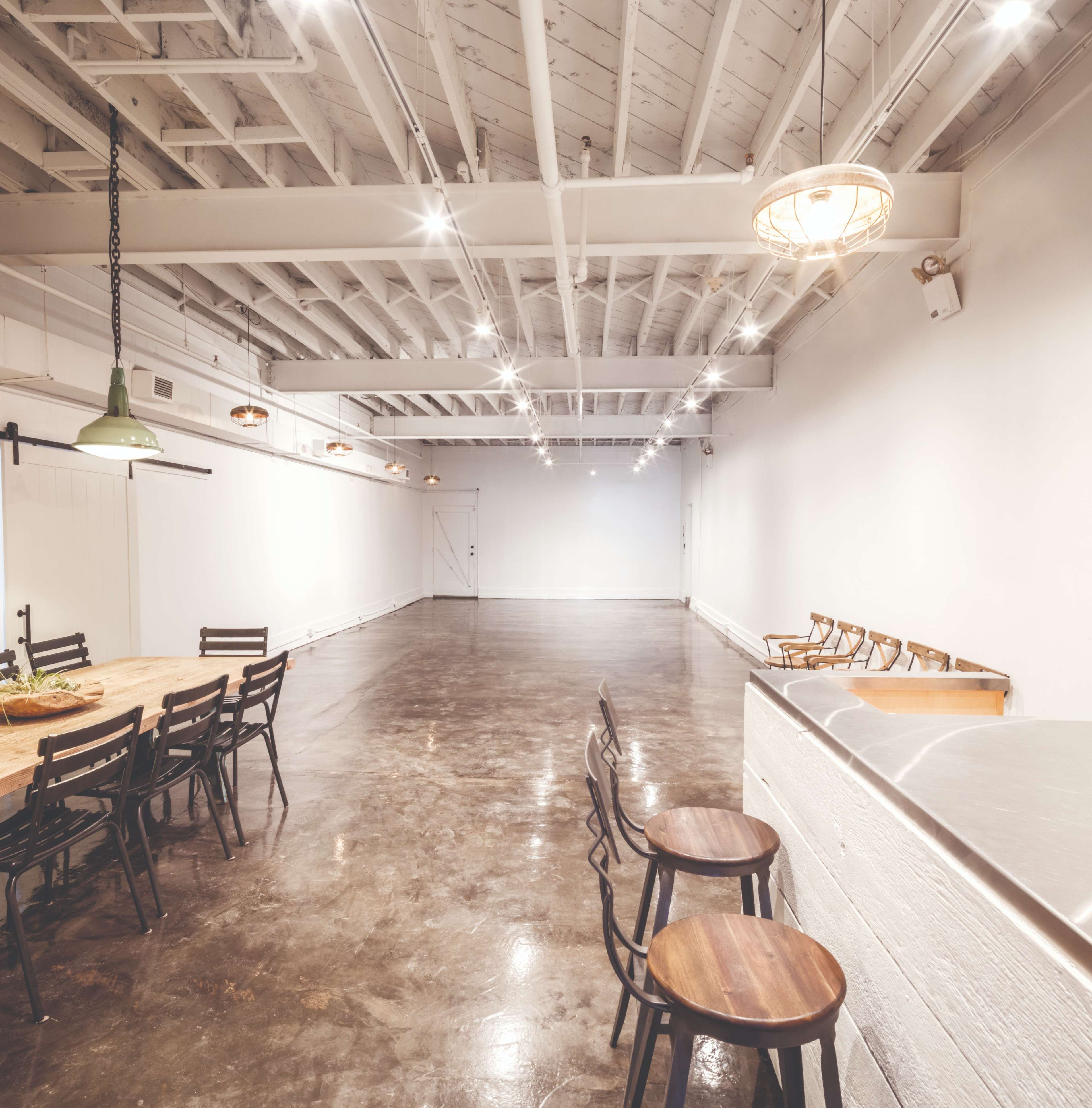 The image shows a spacious, minimalist interior with polished concrete floors, a long wooden table surrounded by chairs, and soft lighting from hanging fixtures.