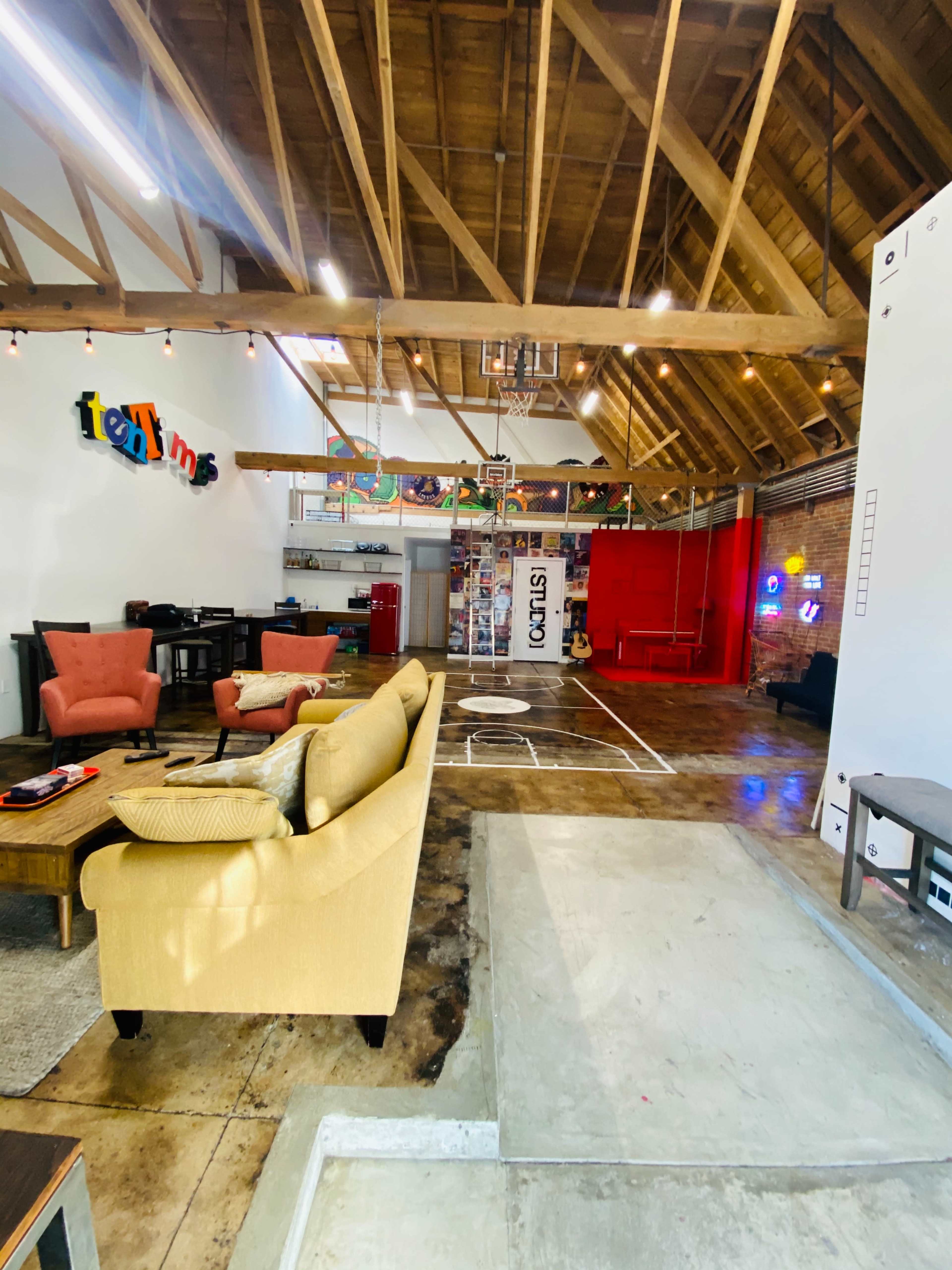 A spacious, well-lit loft with high wooden ceilings, featuring a mix of casual seating areas, a basketball hoop, and an exposed brick wall.