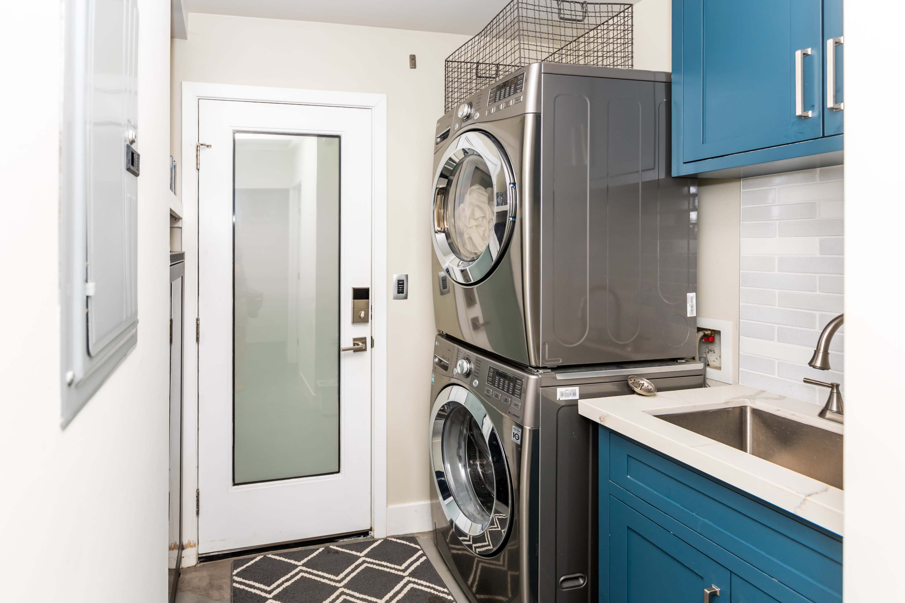 A laundry area with stacked washer and dryer units beside a sink, having blue cabinetry and a glass-paneled door.