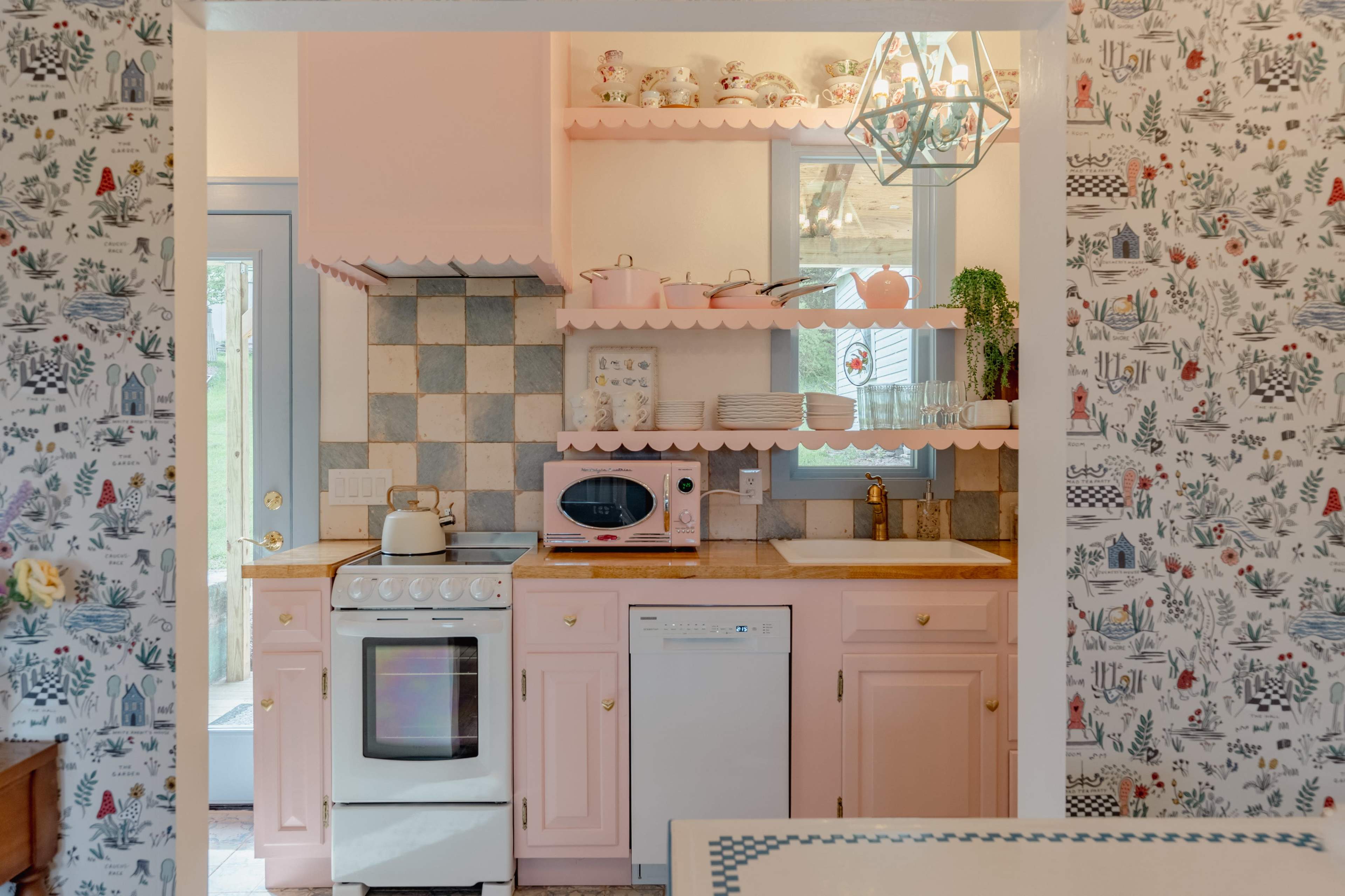 The image shows a pastel pink kitchen featuring a stove, microwave, and dishwasher, alongside decorative shelving and patterned wallpaper.