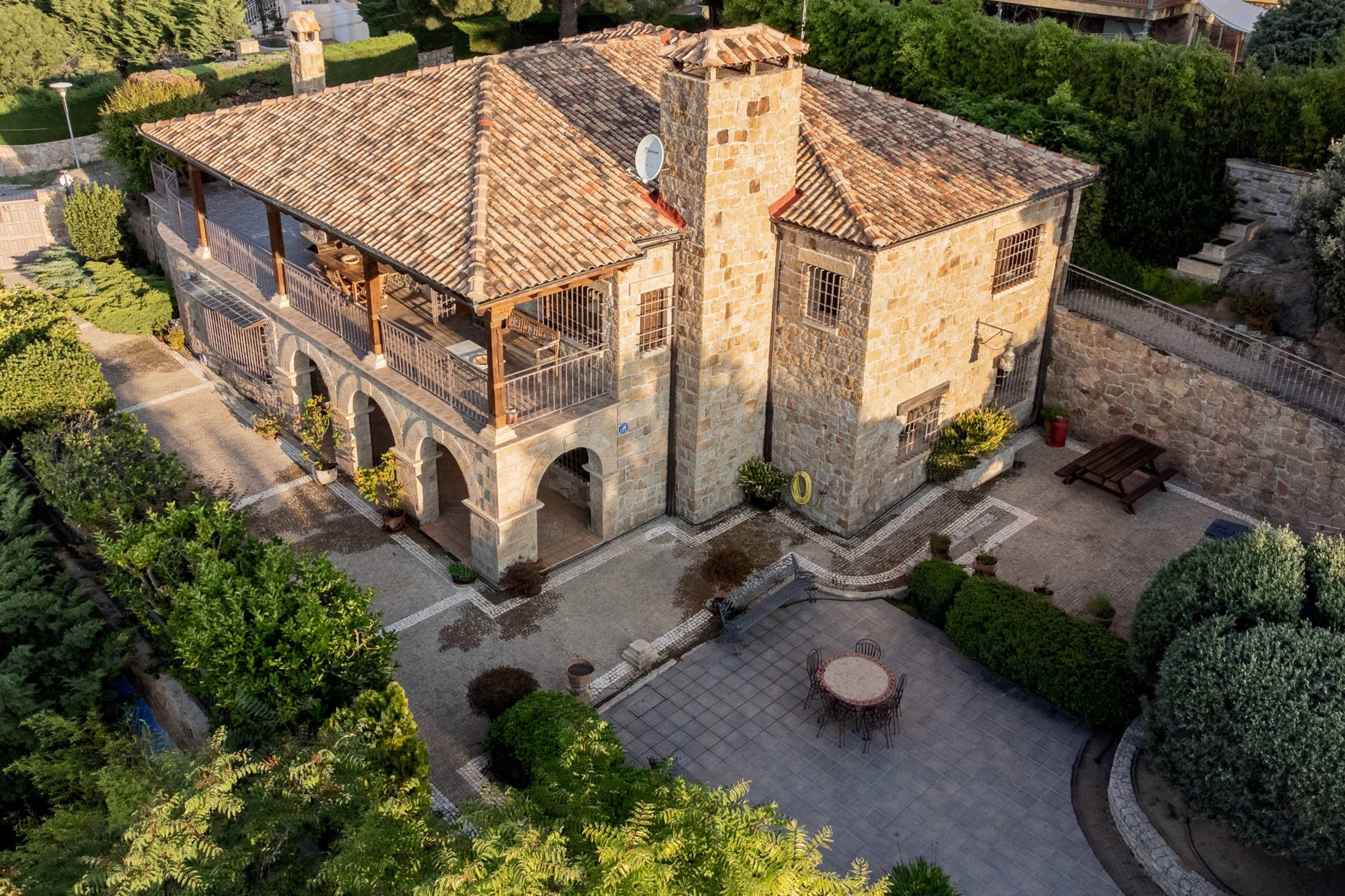 A stone house with a tiled roof, featuring a terrace with arches and surrounded by well-maintained landscaping.