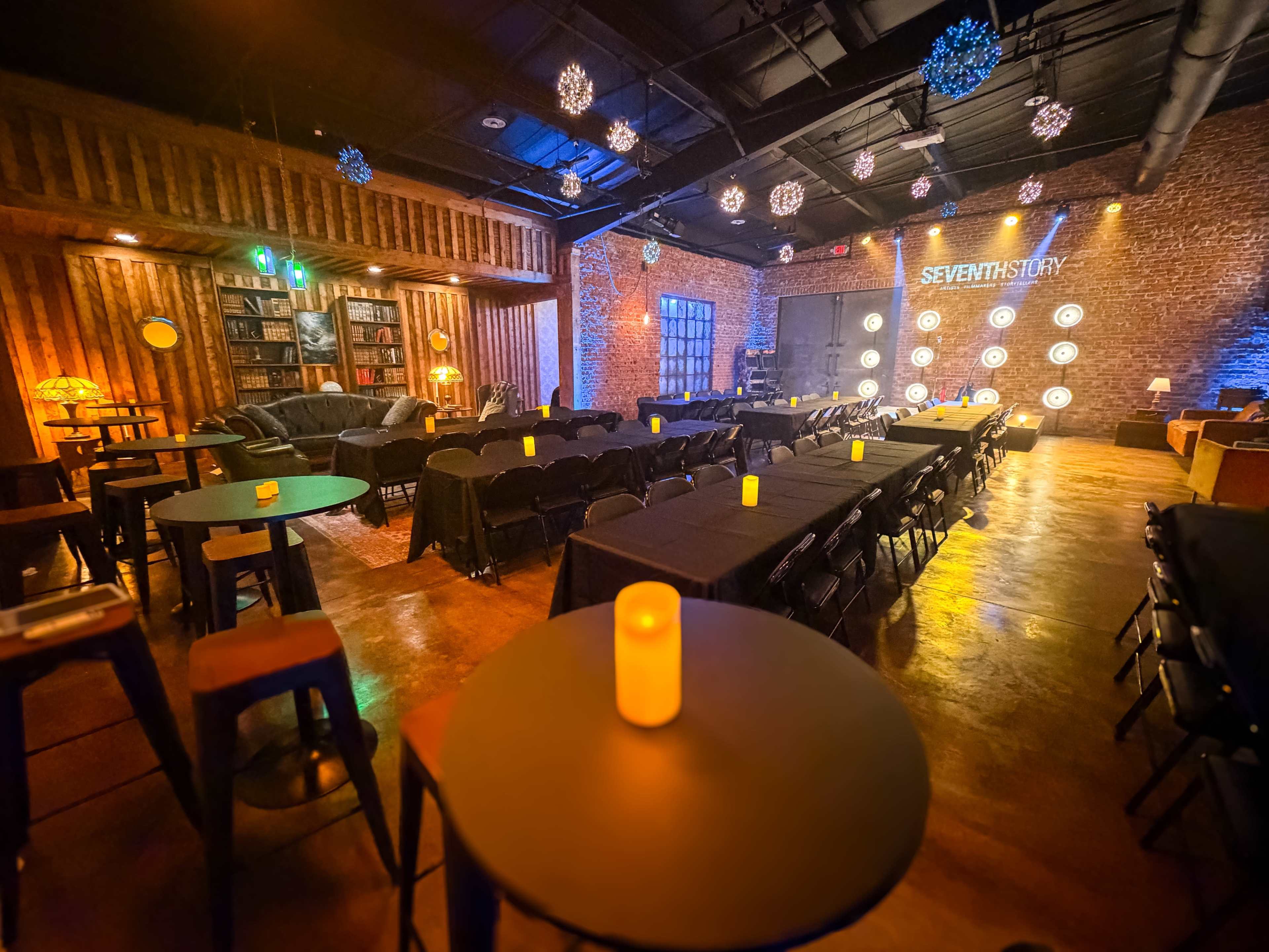 The image shows an indoor event space featuring rows of tables and chairs arranged for an audience, illuminated by soft lighting and decorative elements.