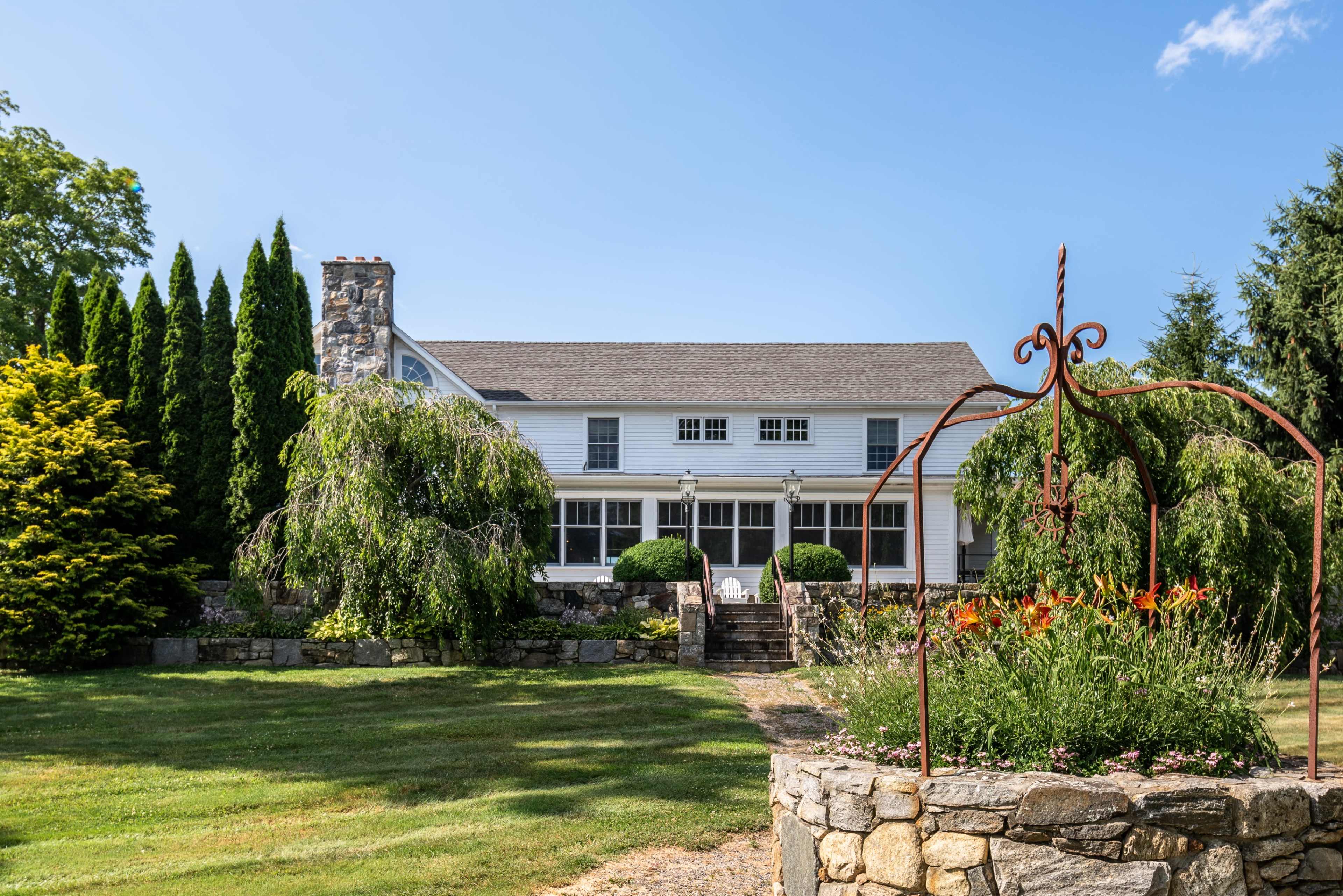 A large white house with a stone chimney is surrounded by lush greenery and features a stone pathway leading to a welcoming front porch.
