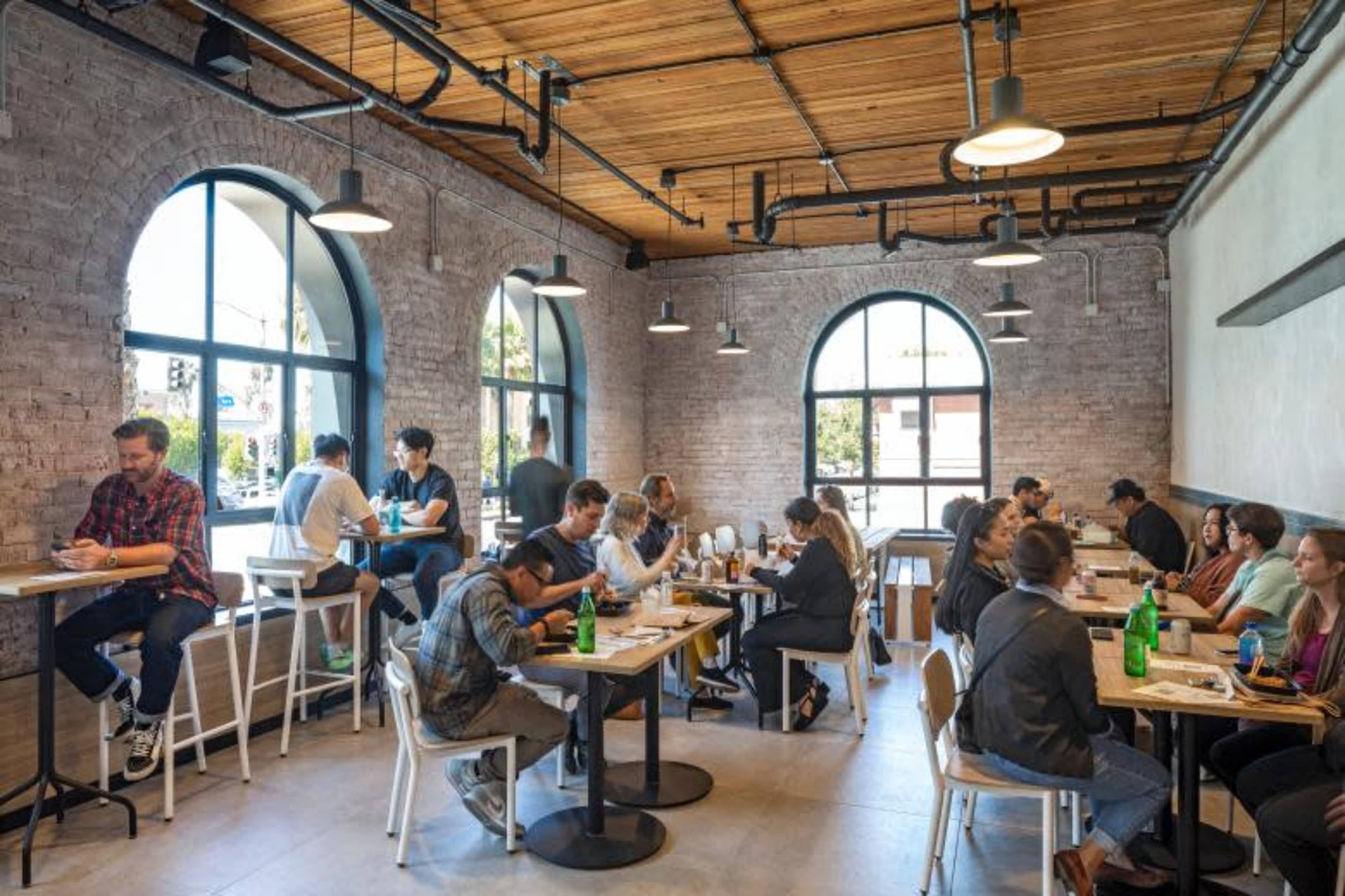 A busy restaurant interior filled with diners seated at tables and interacting in a modern space with large windows and exposed brick walls.