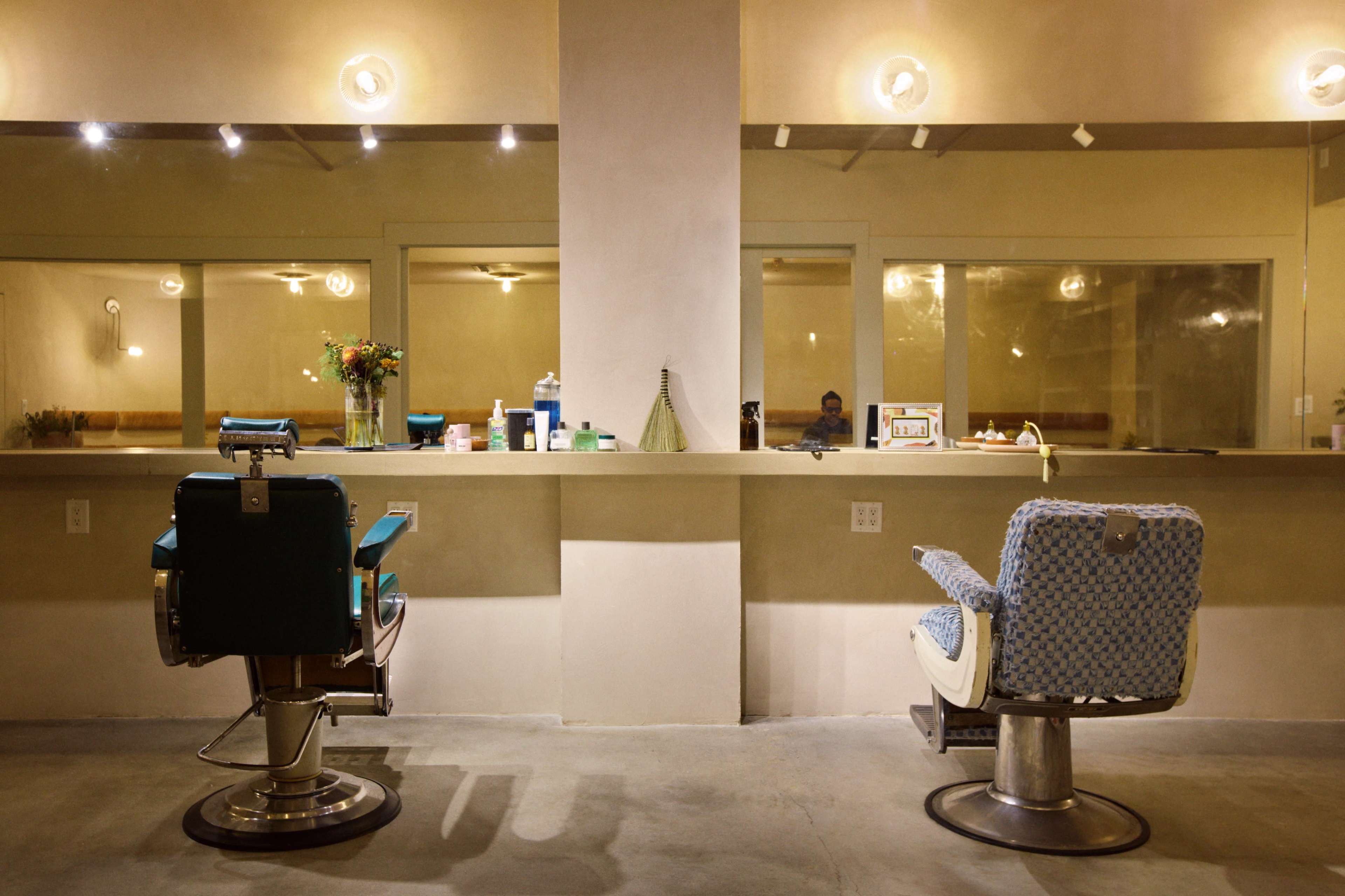 The image shows a minimalist salon interior featuring two styling chairs positioned in front of a mirror and a service counter.
