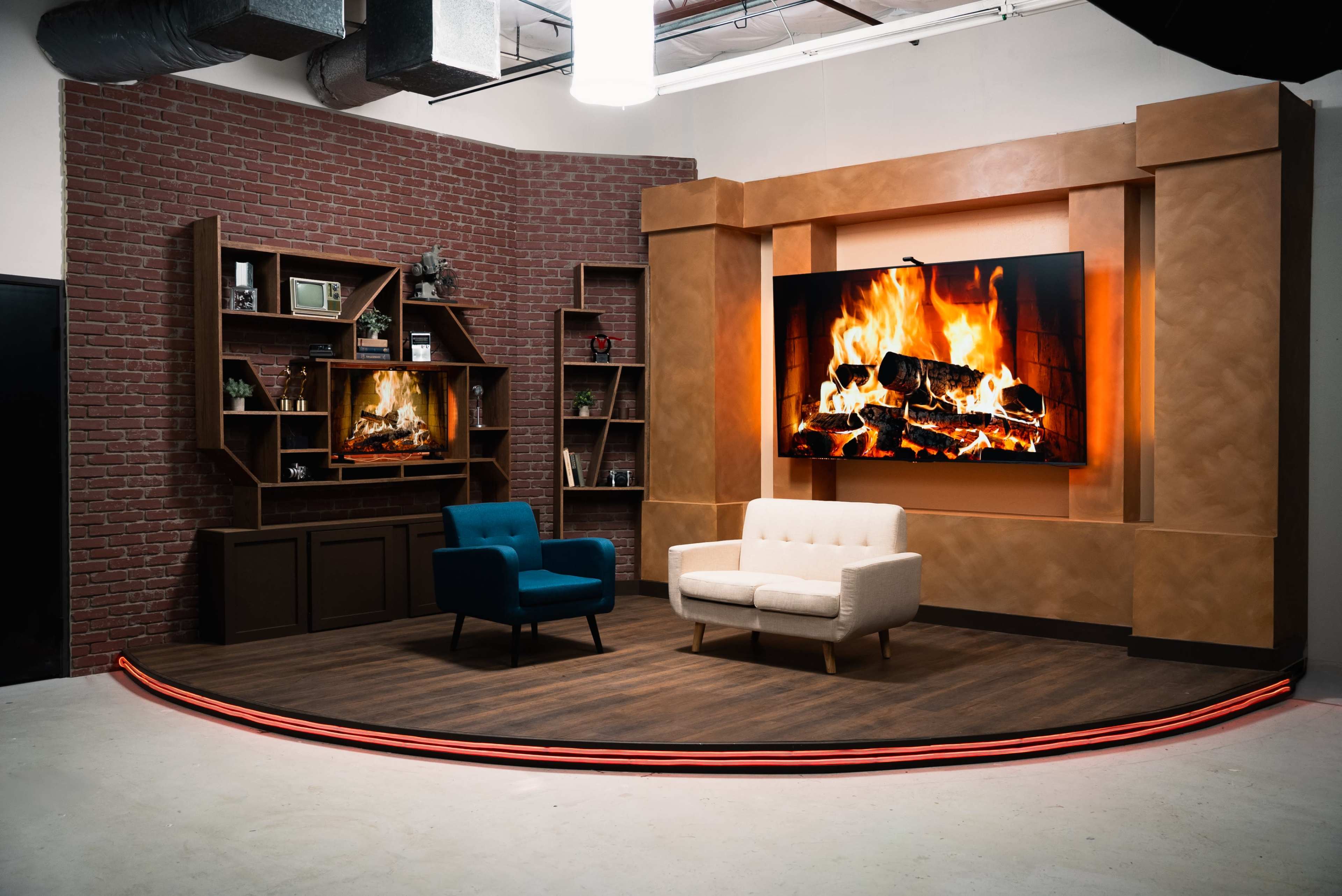 The image depicts a modern studio set with a fireplace display, two chairs, and a brick-accented wall featuring shelves.