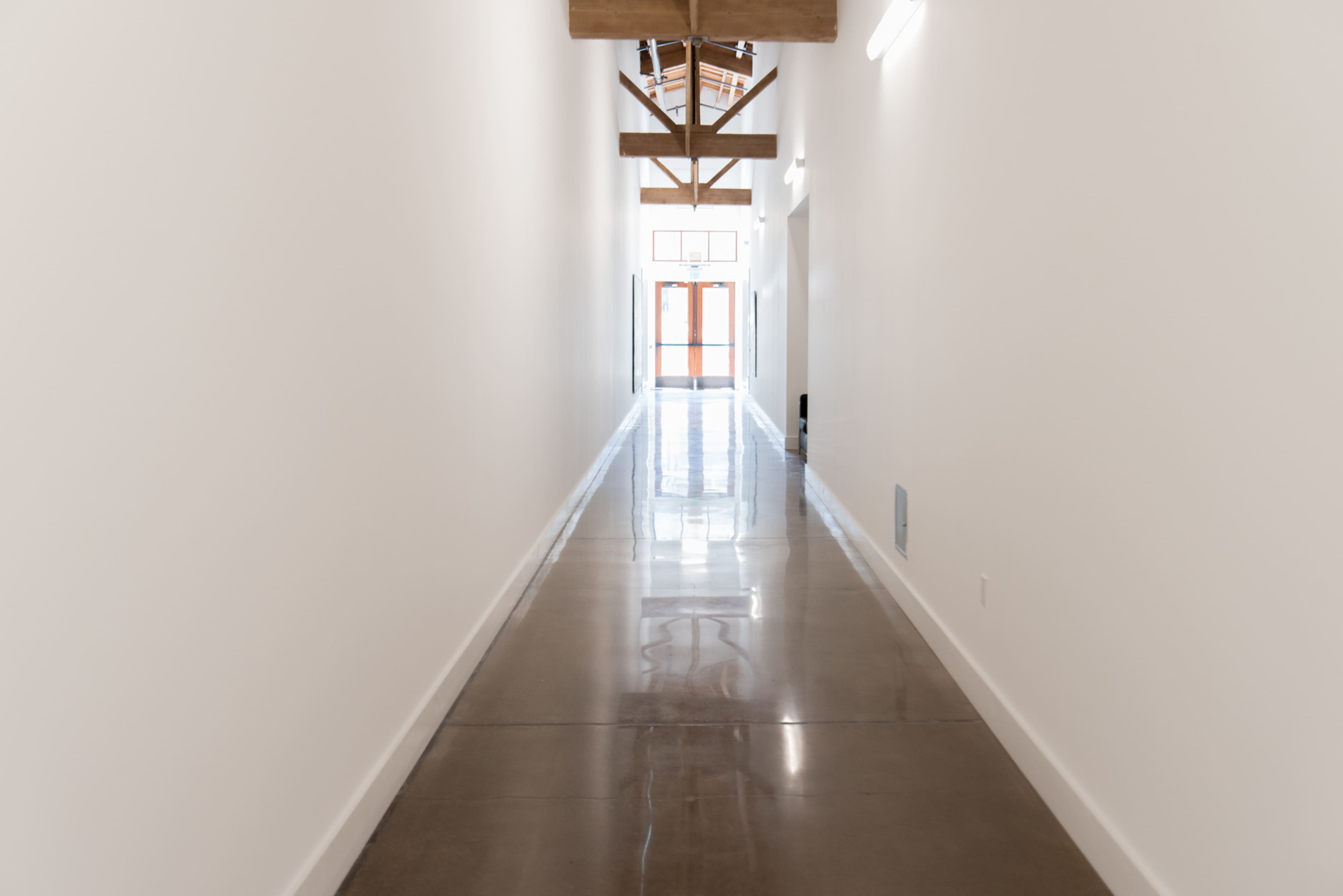 A long, narrow hallway with polished concrete floors and wooden beams overhead leads to a set of double doors at the end.