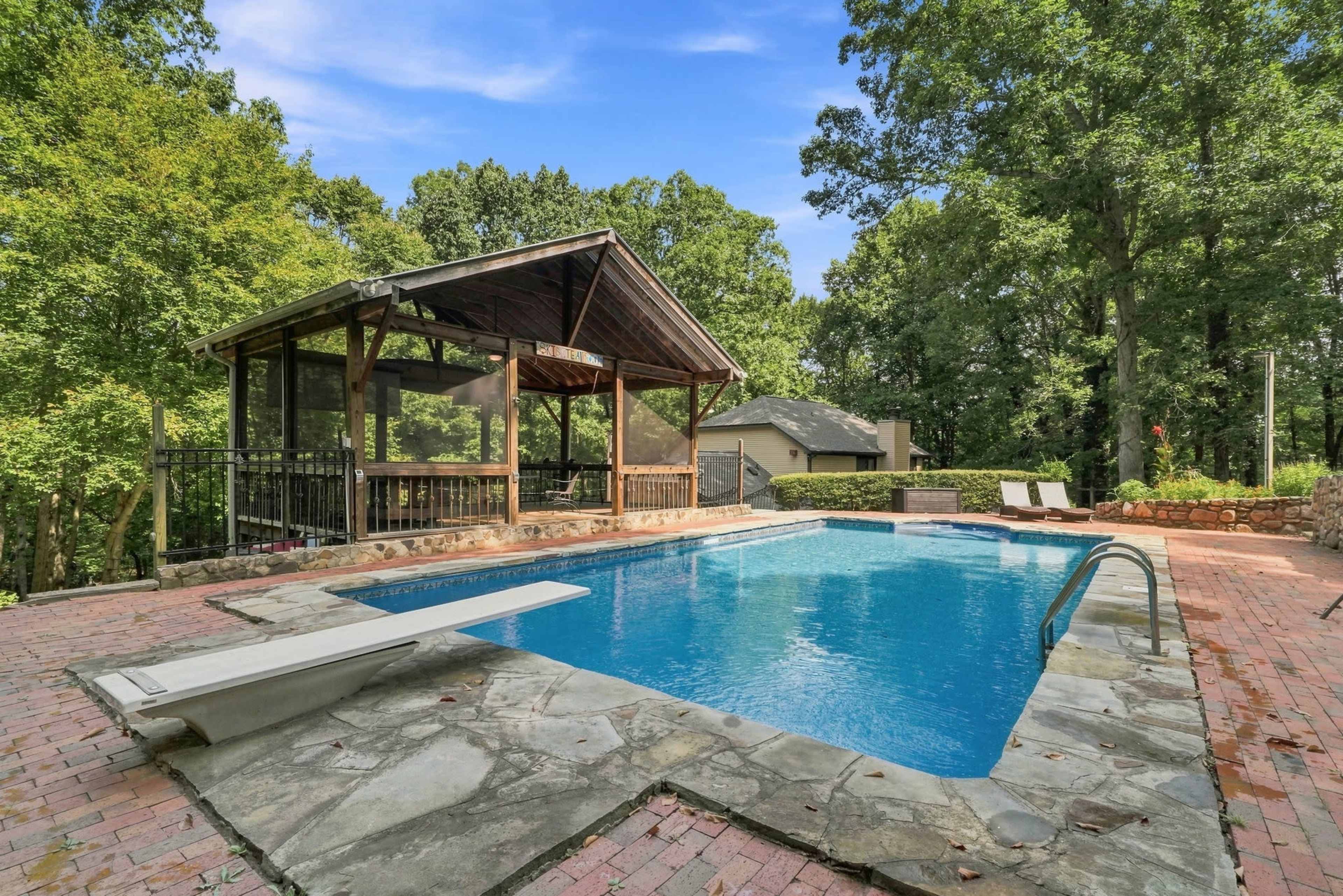 The image shows a rectangular swimming pool with a diving board, surrounded by a stone patio and a wooden gazebo in a wooded backyard.