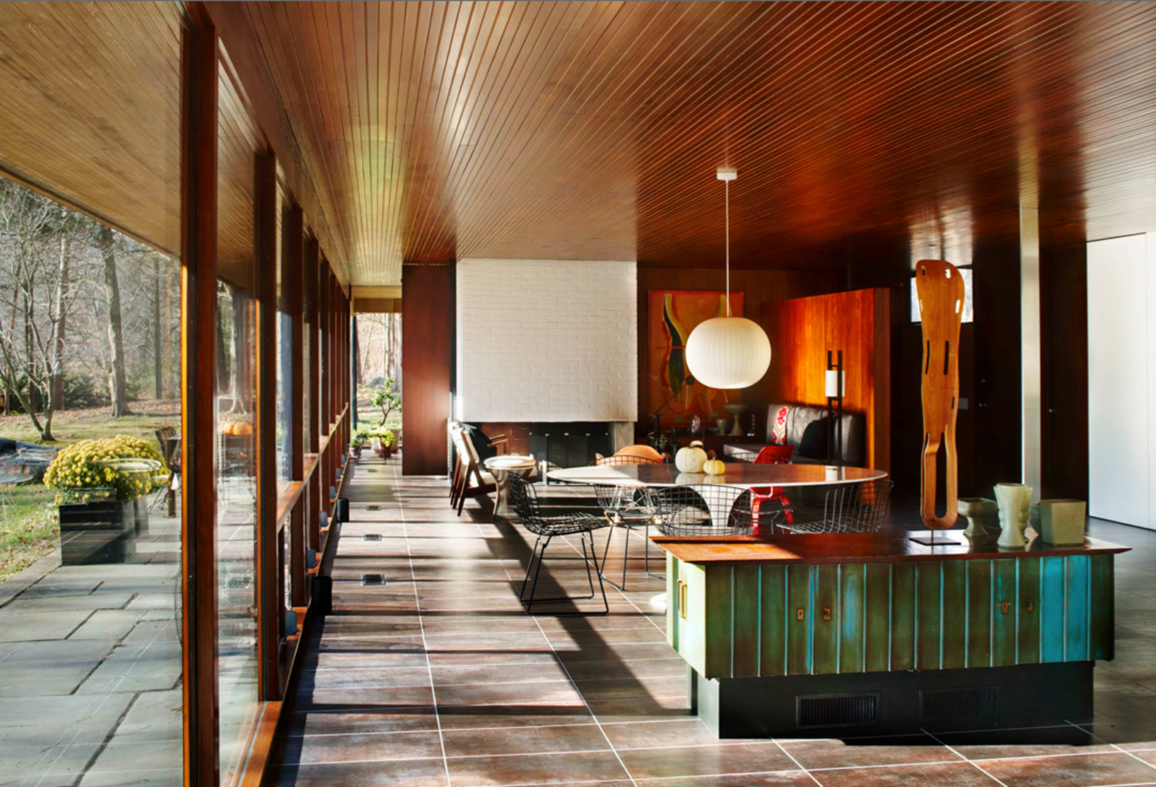 The image shows a modern interior space featuring a wooden ceiling, large glass windows, a dining area with a circular table, and a cozy sitting arrangement near a fireplace.