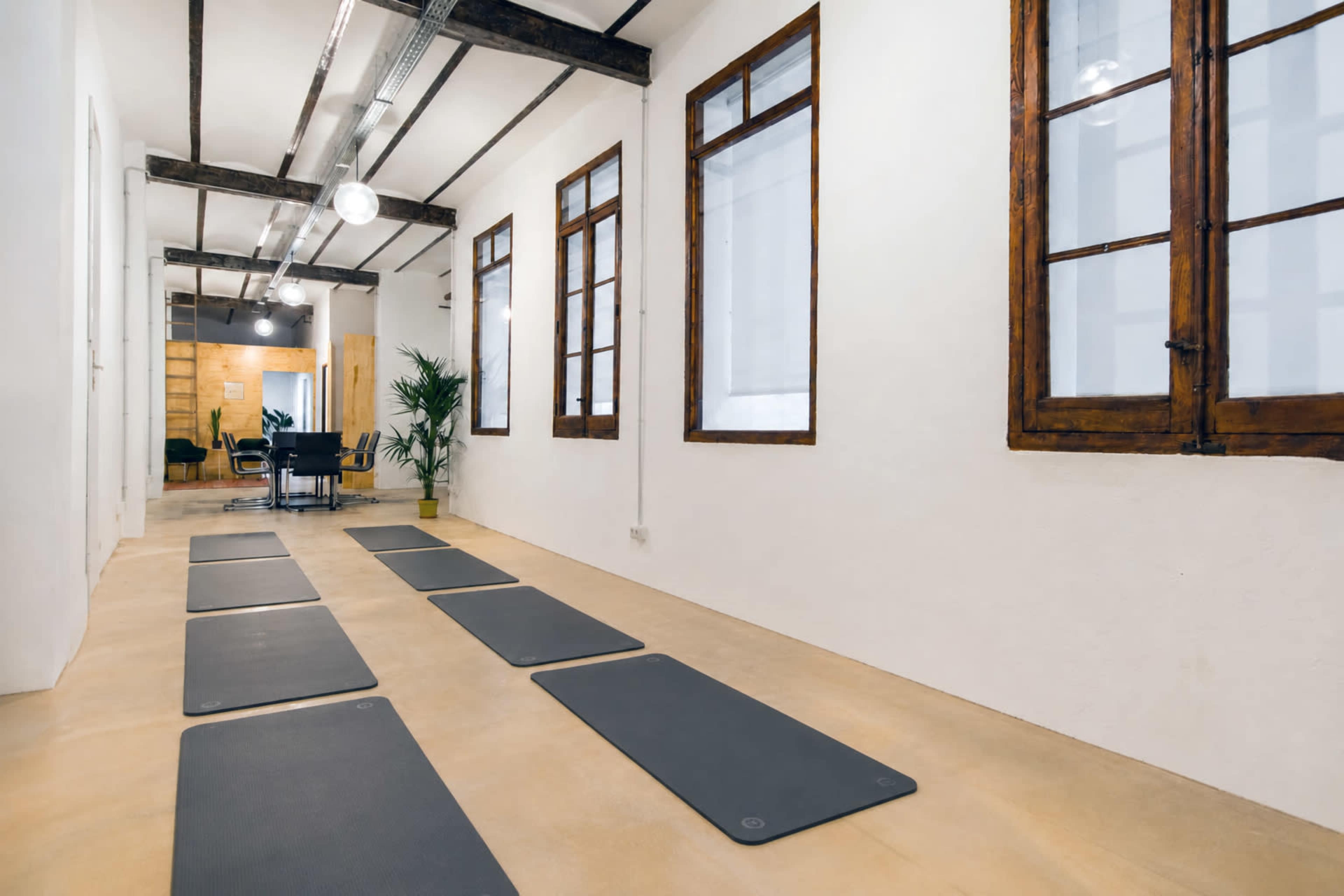 The image shows a bright room with wooden beams, featuring several yoga mats lined up on a smooth floor and large windows allowing natural light to enter.