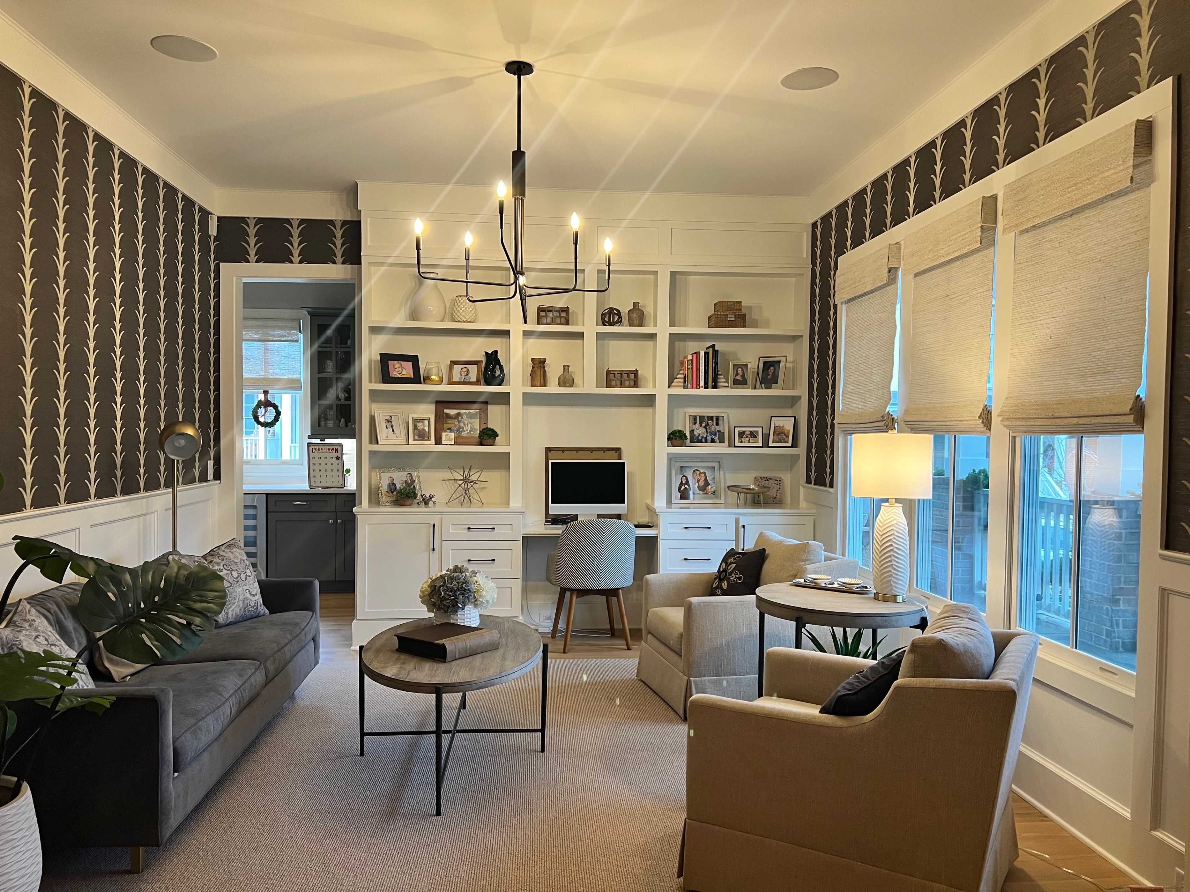A cozy home office with two sofas, a circular coffee table, a bookshelf filled with decorative items, and a desk with a computer, all set against a patterned wallpaper.