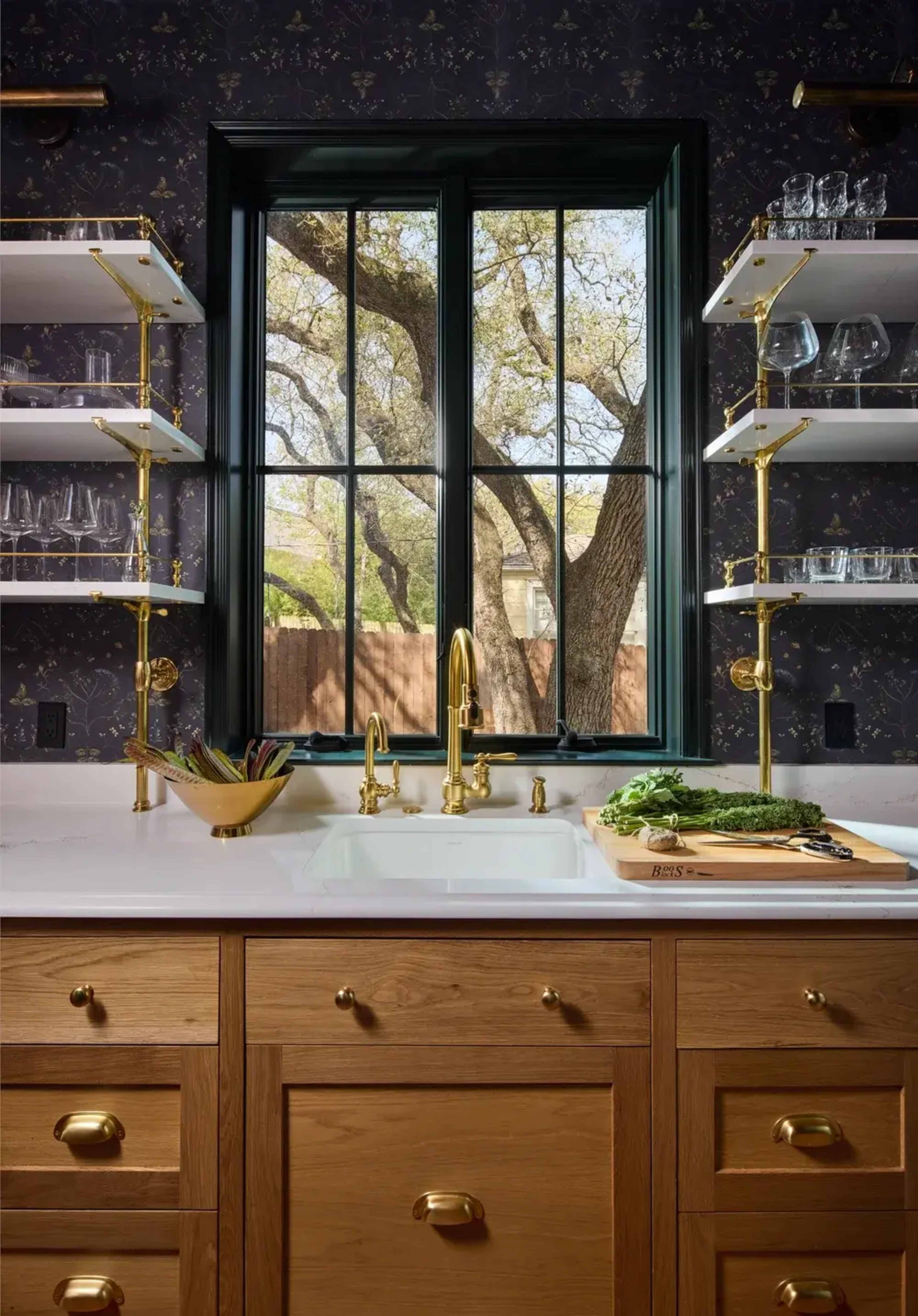 A modern kitchen features a window with a black frame overlooking a tree, with wooden cabinetry and open shelves displaying glassware.