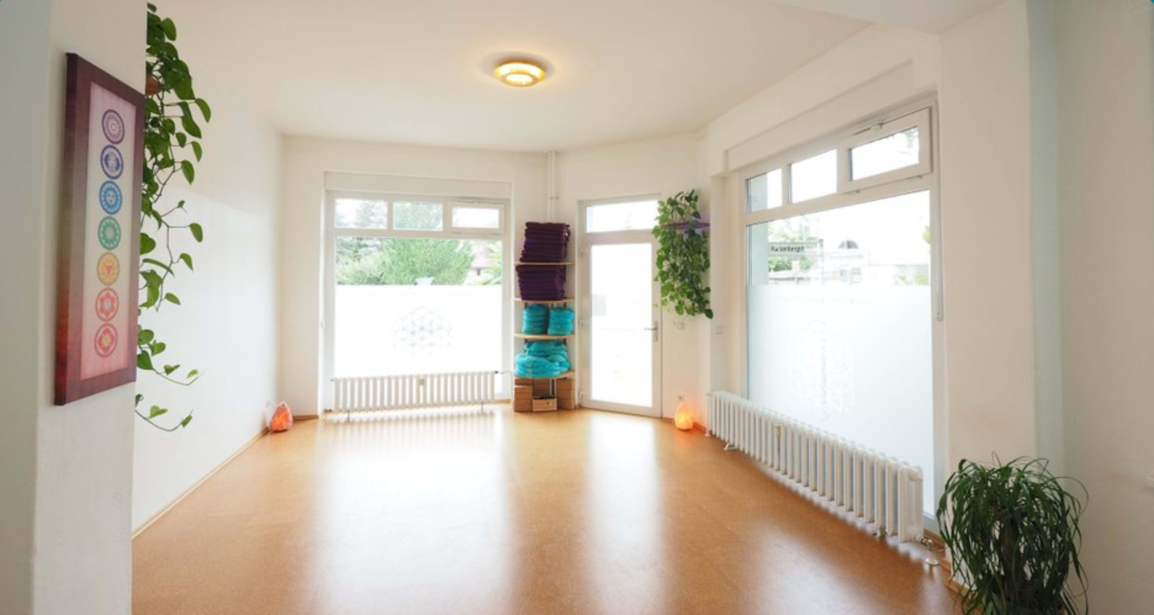 A bright, empty room with large windows, natural light, and plants, featuring a wooden floor and stacked yoga mats.