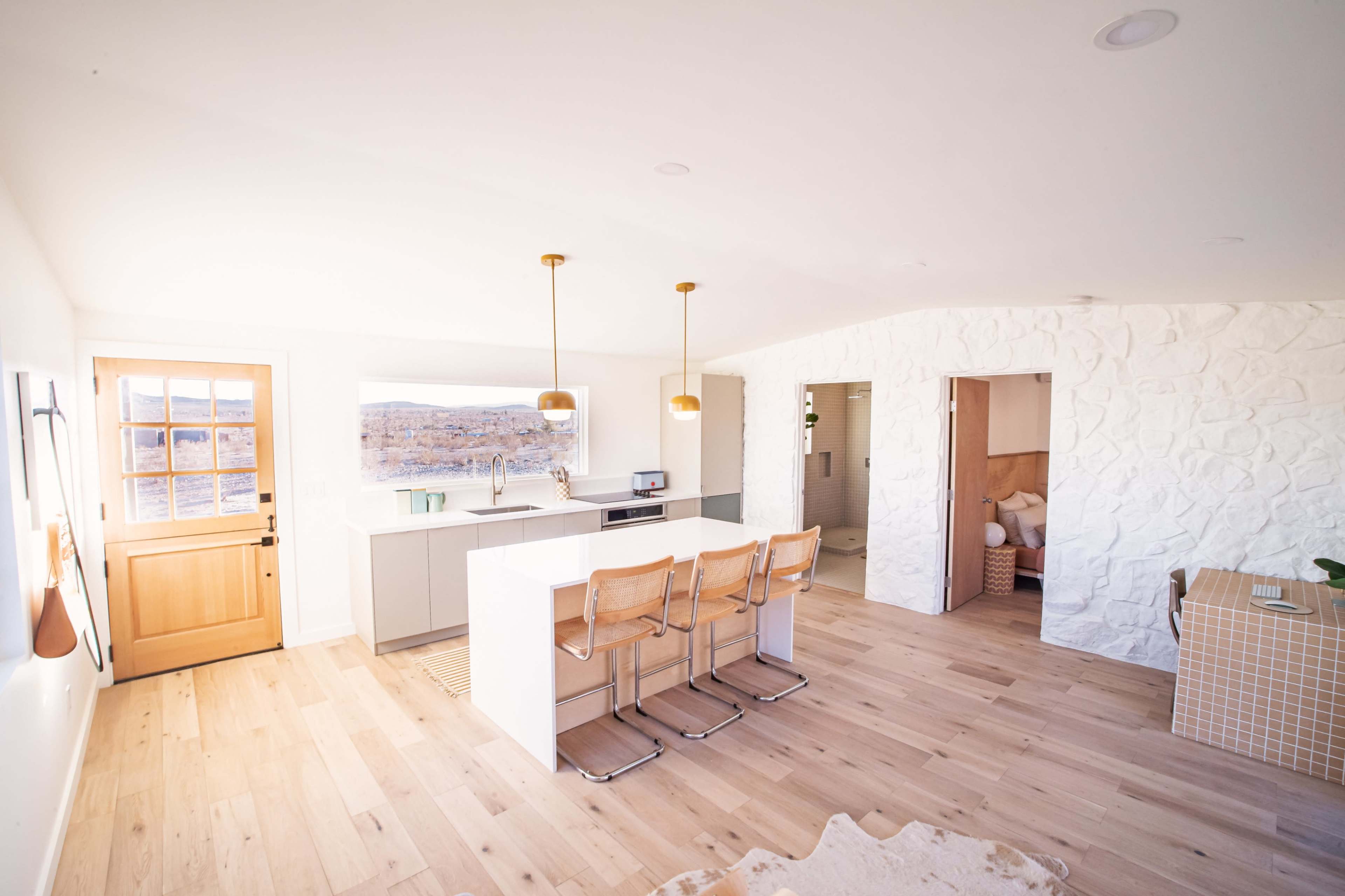 A modern kitchen and living space featuring light wood flooring, a large island with bar stools, and a bright, minimalist design.