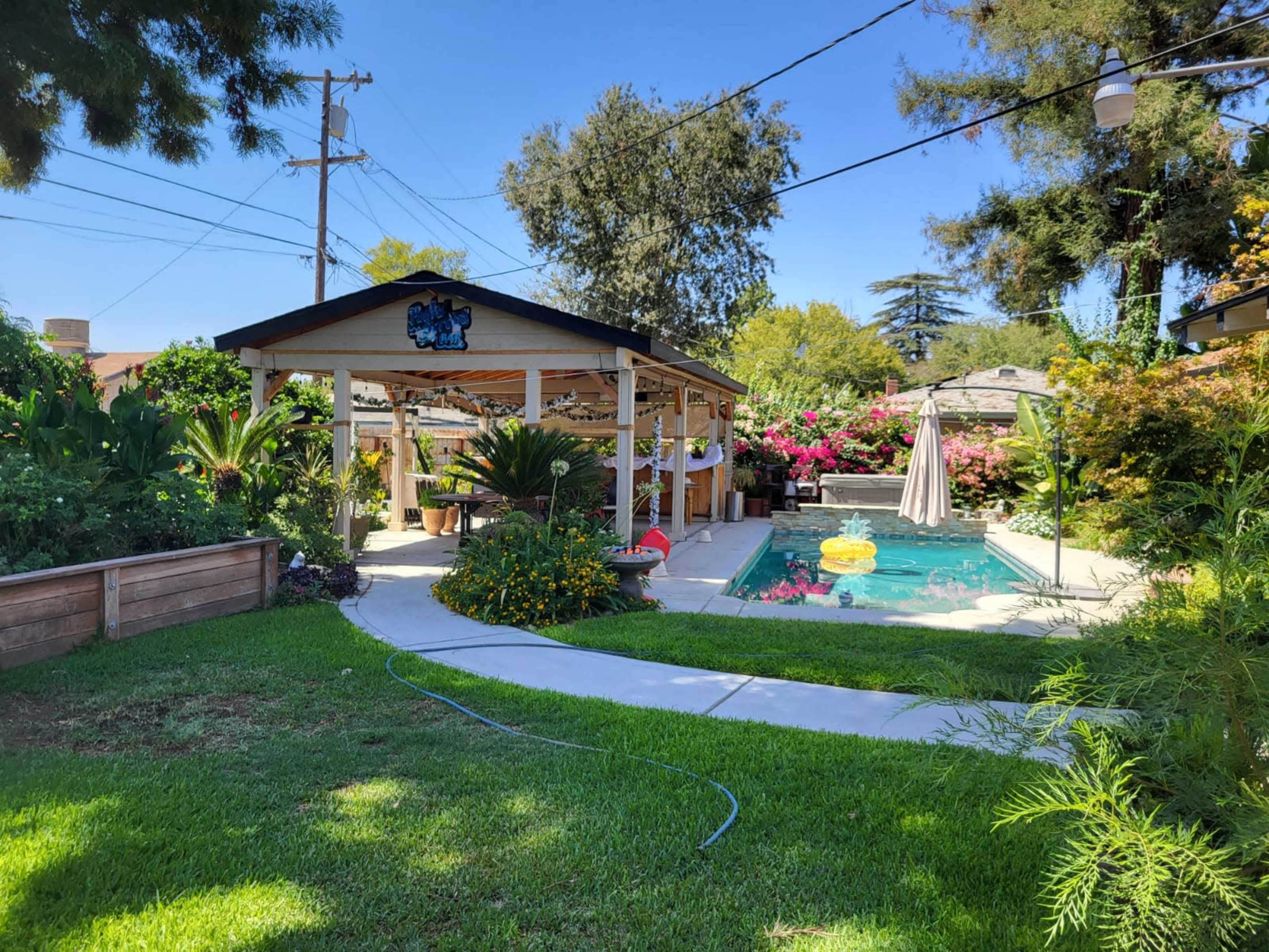 A well-maintained backyard features a swimming pool surrounded by lush plants and a covered patio with seating.