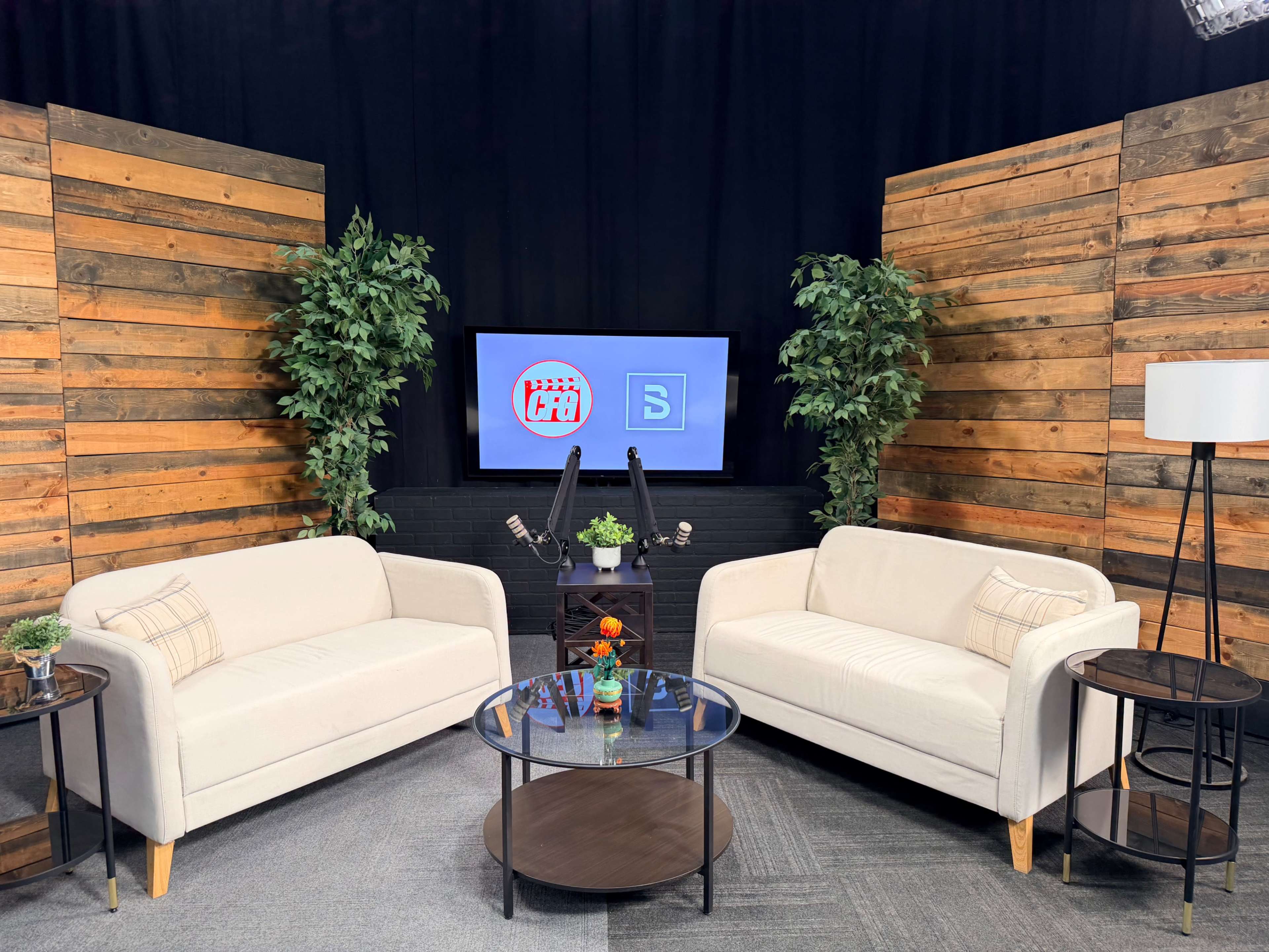 The image shows a well-lit studio setting with two white sofas facing a circular glass table, flanked by wooden paneling and potted plants, featuring a monitor displaying logos in the background.