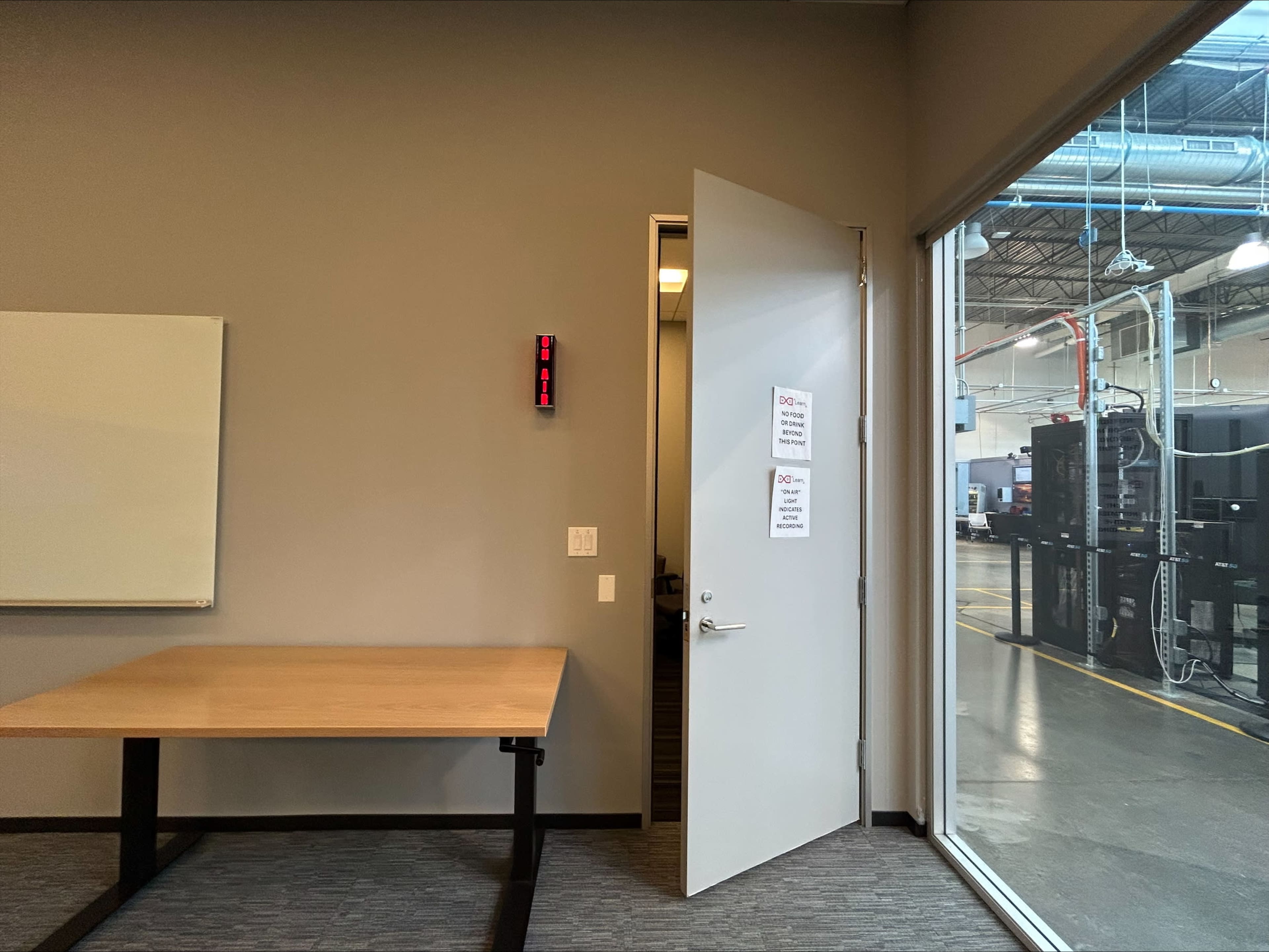 The image shows a partially open door leading to a warehouse area with industrial equipment, alongside a table and a wall-mounted digital clock displaying the time.