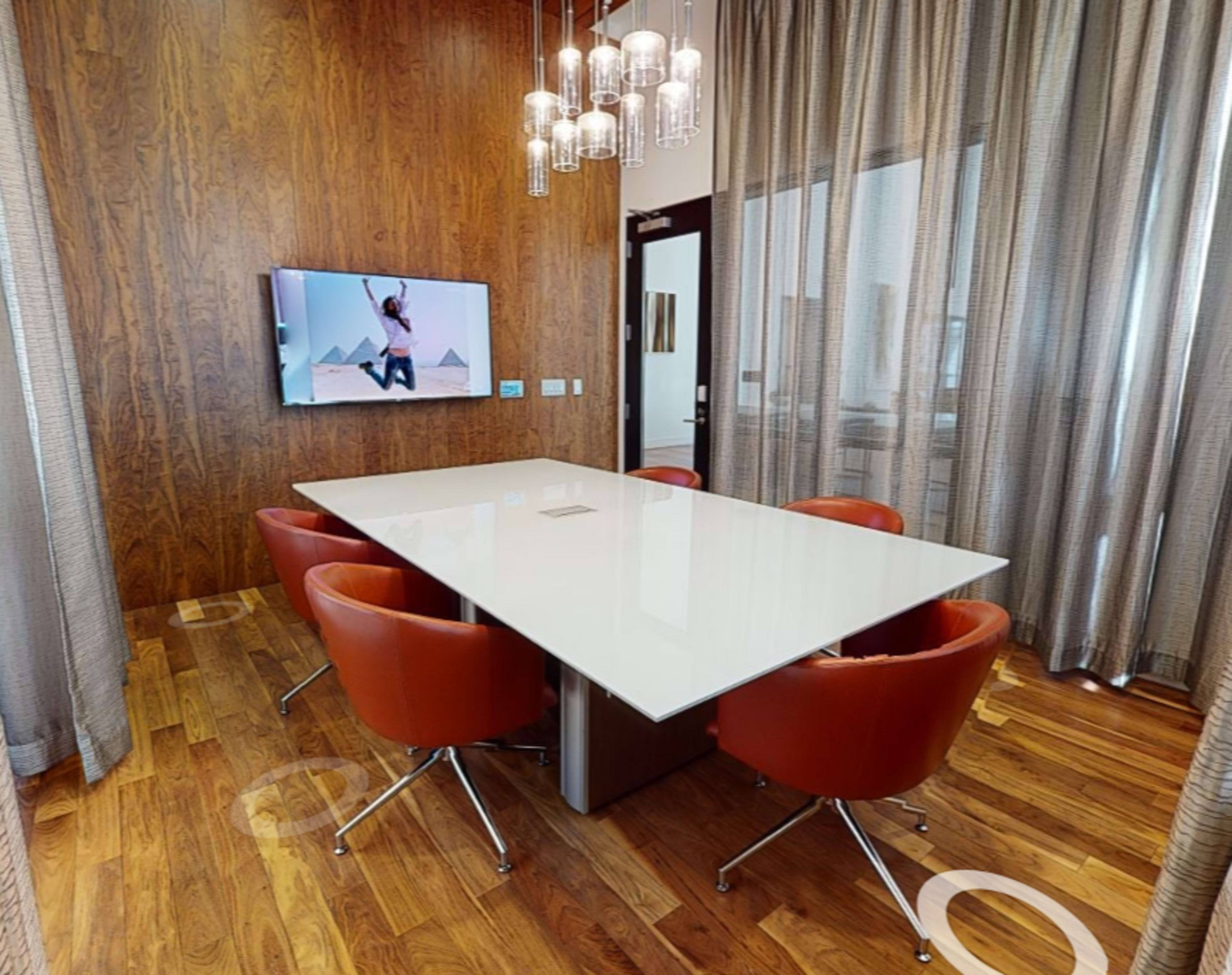 A modern conference room features a white table surrounded by four red leather chairs, with a large TV displaying a scenic image on the wood-paneled wall.