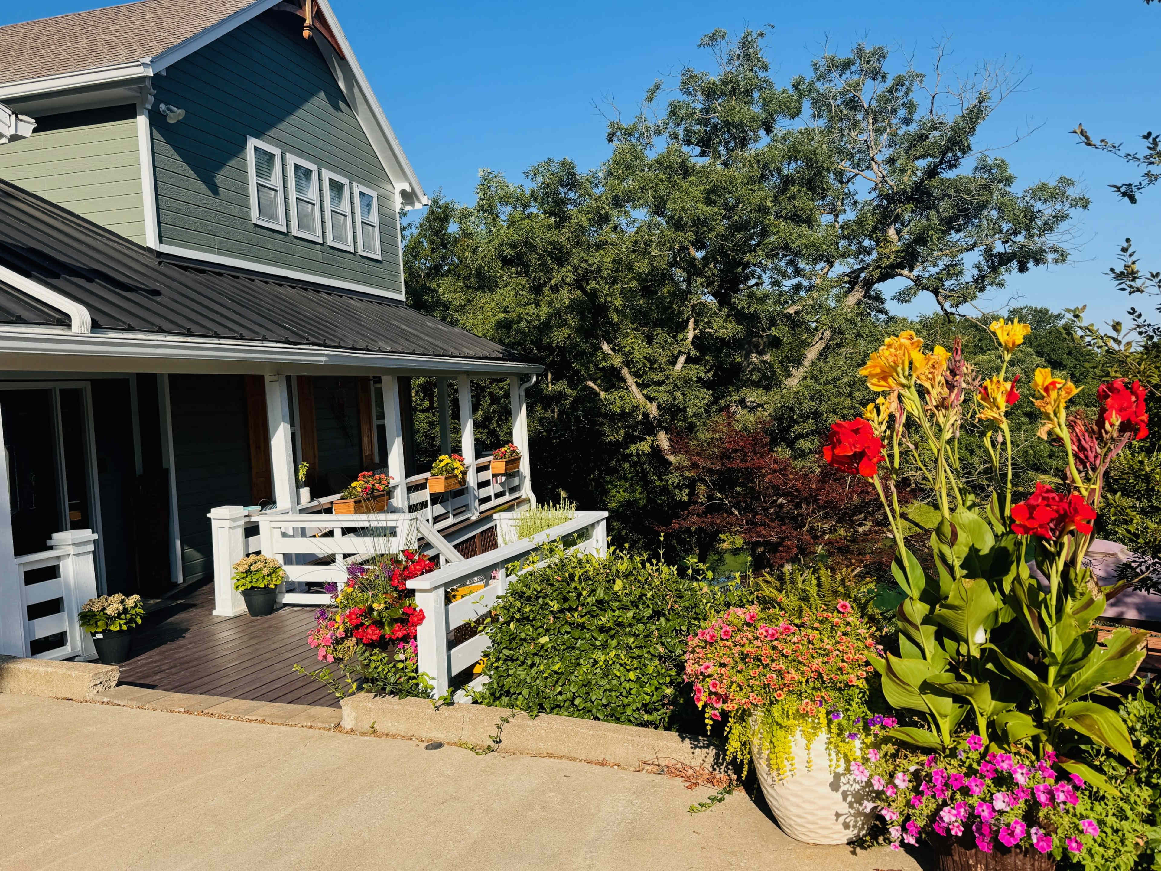 A two-story green house with a black roof features a front porch surrounded by flowering plants and colorful pots.